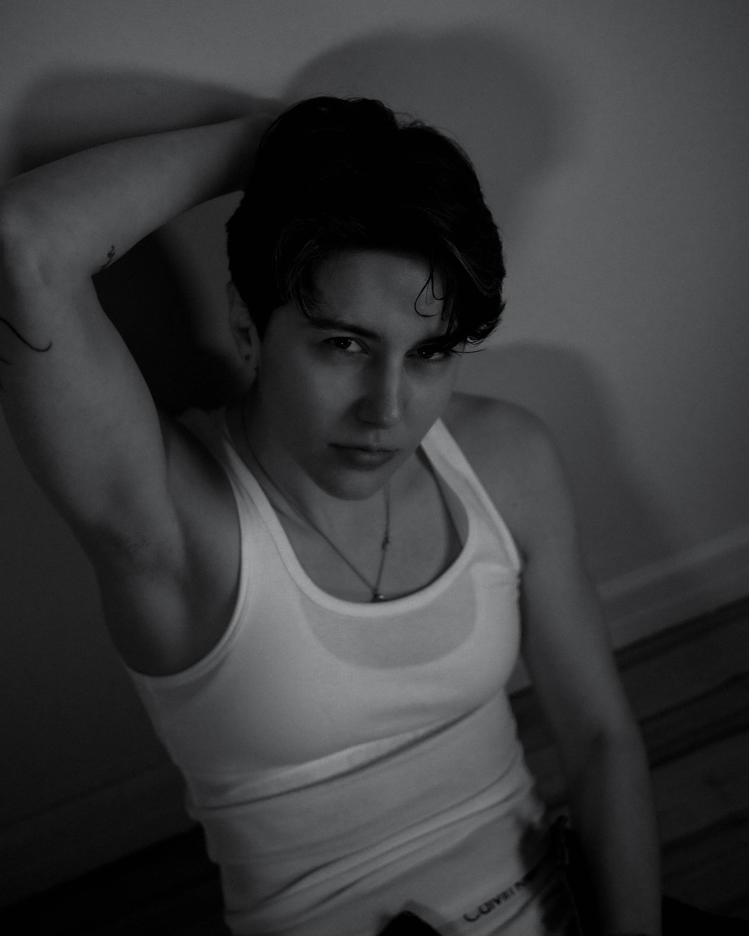 Jeune homme à court noir, portant un tank top blanc, posant avec bras levé derrière la tête, regardant l'objectif.