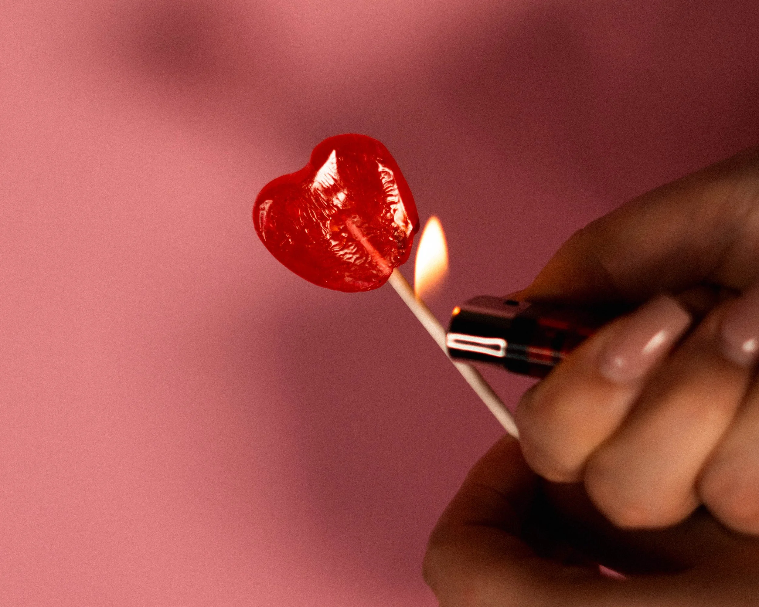 Une main tenant un bâtonnet avec une sucette en forme de cœur rouge brillant, avec un filtre à briquet allumé en dessous.