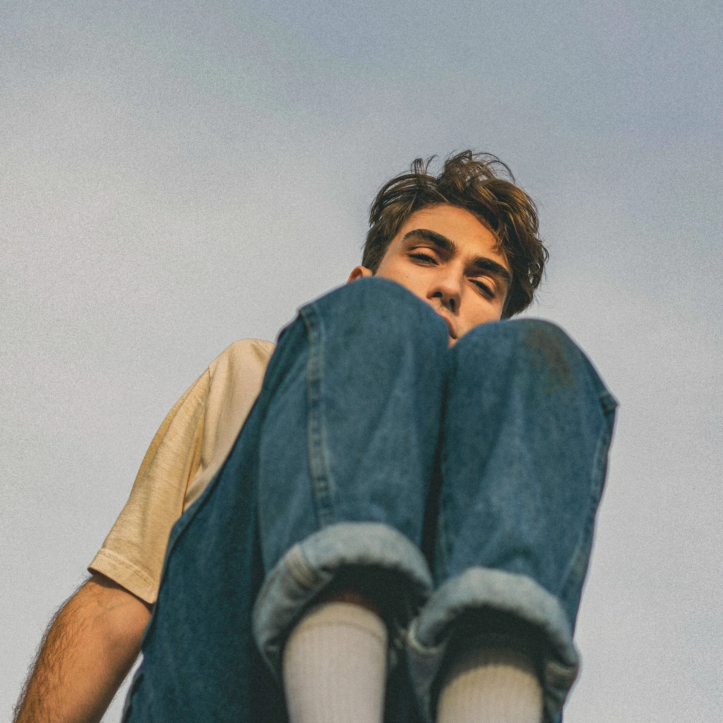 Jeune homme aux cheveux bruns, portant un jean, souriant, vu de près avec un ciel clair en arrière-plan.
