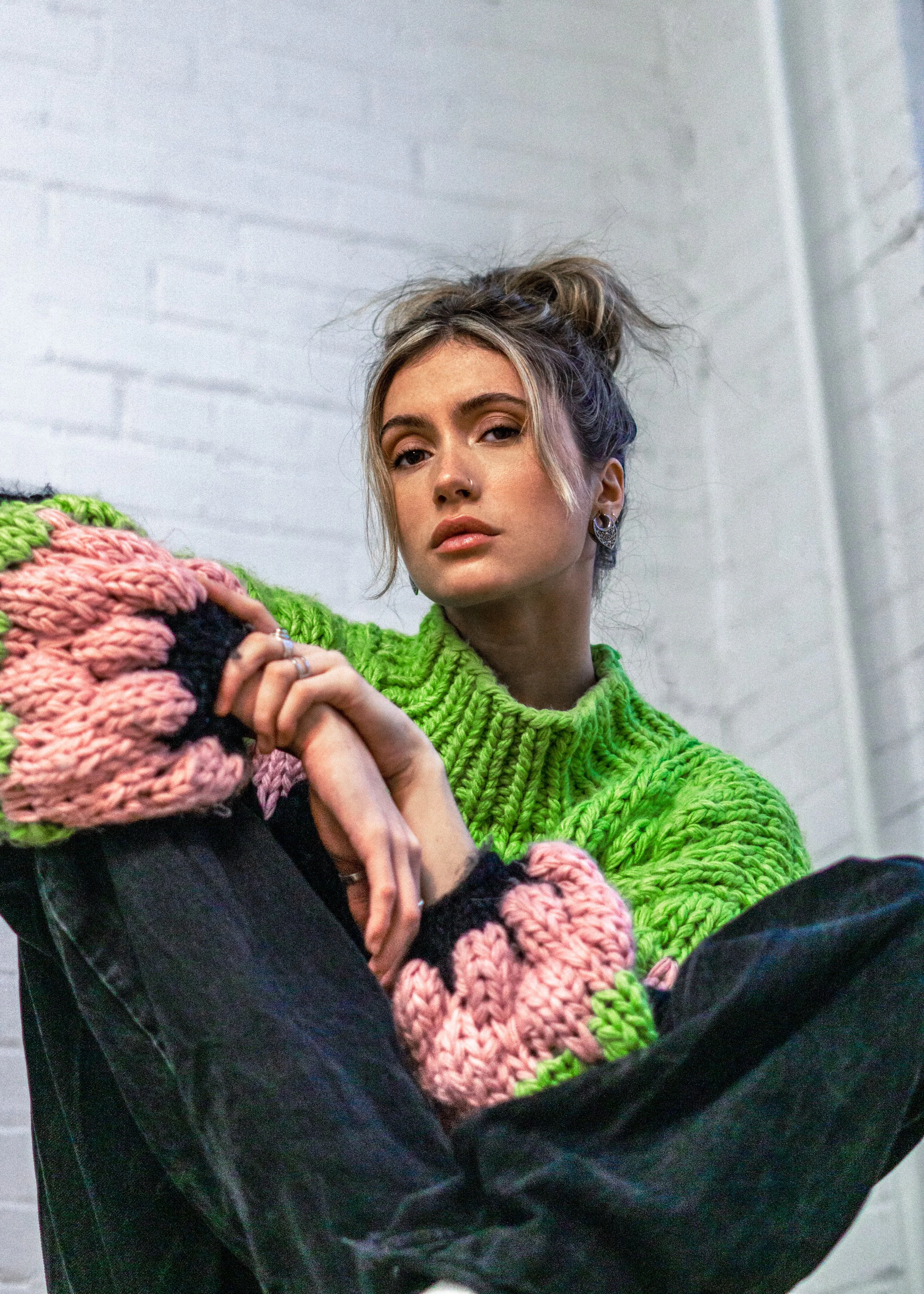 Une femme portant un pull en laine vert et un vêtement en laine bicolore rose et noir est assise contre un mur blanc en briques, regardant vers l'objectif.