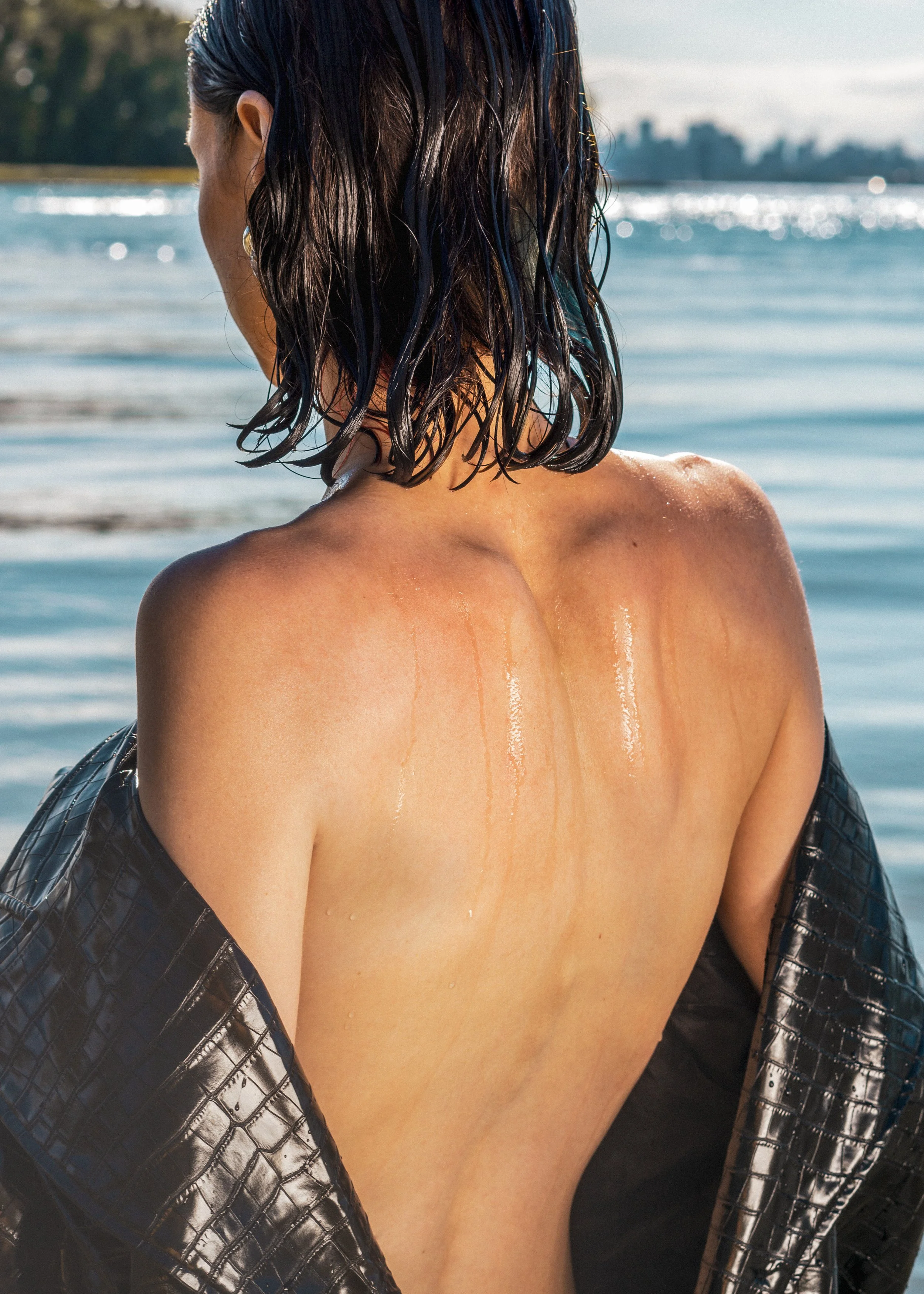 Une femme vue de dos avec des cheveux mouillés, portant une veste en cuir noir, dans un environnement aquatique en bord de mer.
