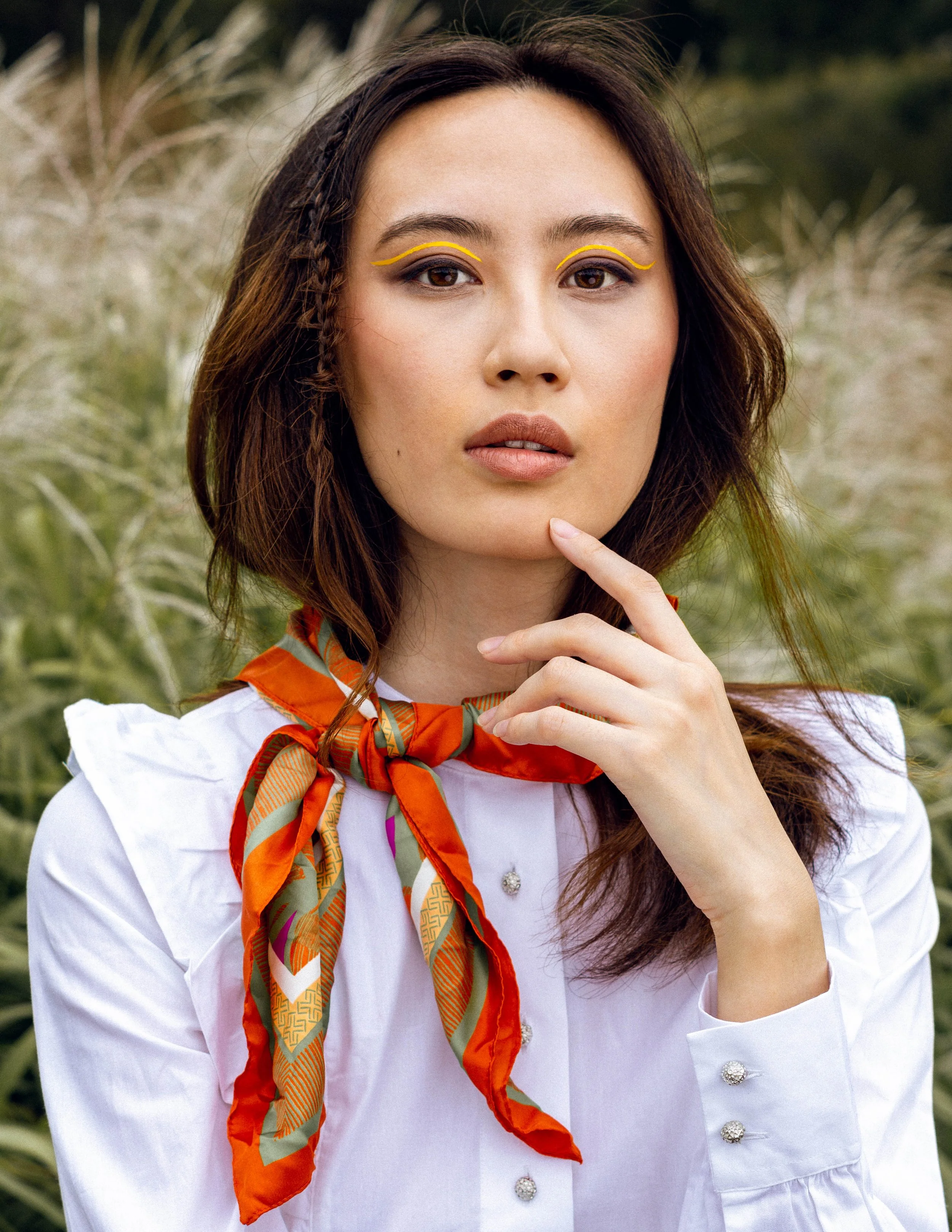 Jeune femme portant un chemisier blanc avec des boutons en forme de pierre, un foulard coloré autour du cou, avec un fond naturel de plantes grasses.