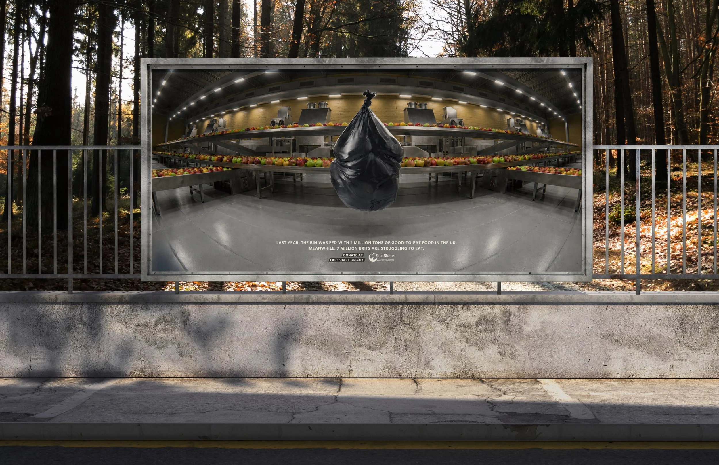 Un cartel publicitario que muestra una imagen de una instalación de producción de alimentos en un bosque, con una caja de recolección llena de frutas. El cartel contiene un texto en la parte inferior que dice: "El año pasado, la caja fue alimentada con 2 millones de toneladas de comida de buen gusto en el Reino Unido. Mientras tanto, 7 millones de británicos luchan por comer." y un enlace para donar.