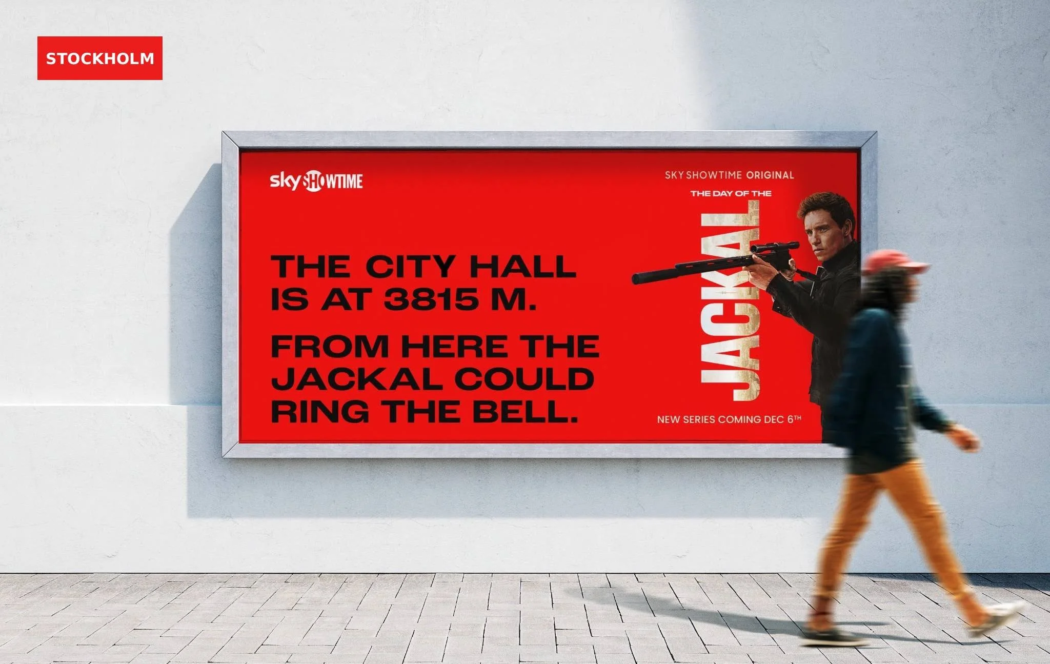 Cartel publicitario en la pared blanca con texto en inglés que dice: "The city hall is at 3815 m. from here the jacks could ring the bell" y una imagen de un hombre con un rifle en un fondo rojo, y una persona que pasa rápidamente delante del cartel.