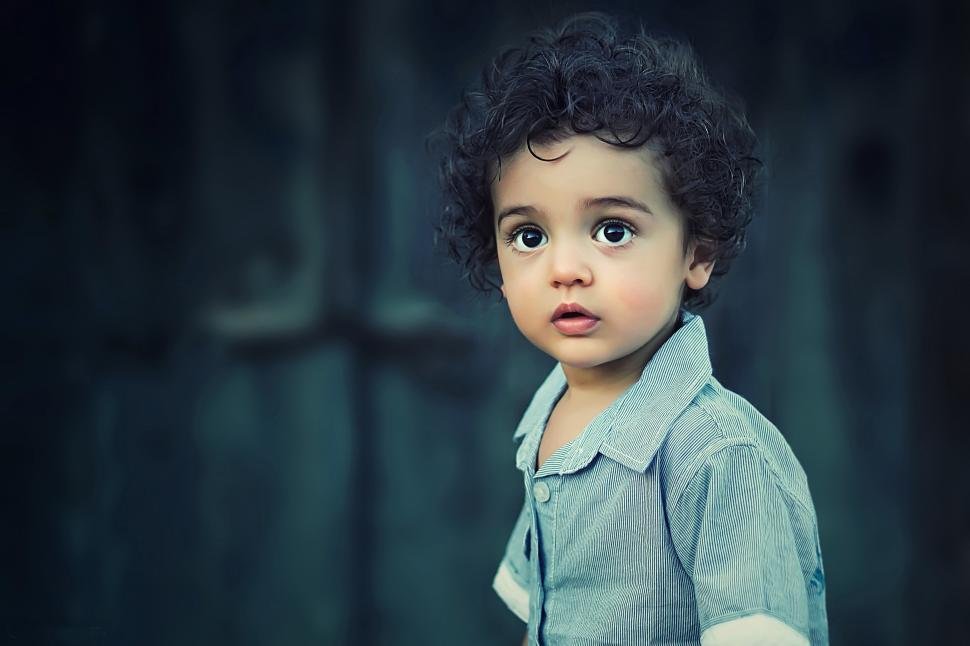 Niño con cabello rizado y ojos grandes, usando una camisa de color gris, en un fondo oscuro y difuso.