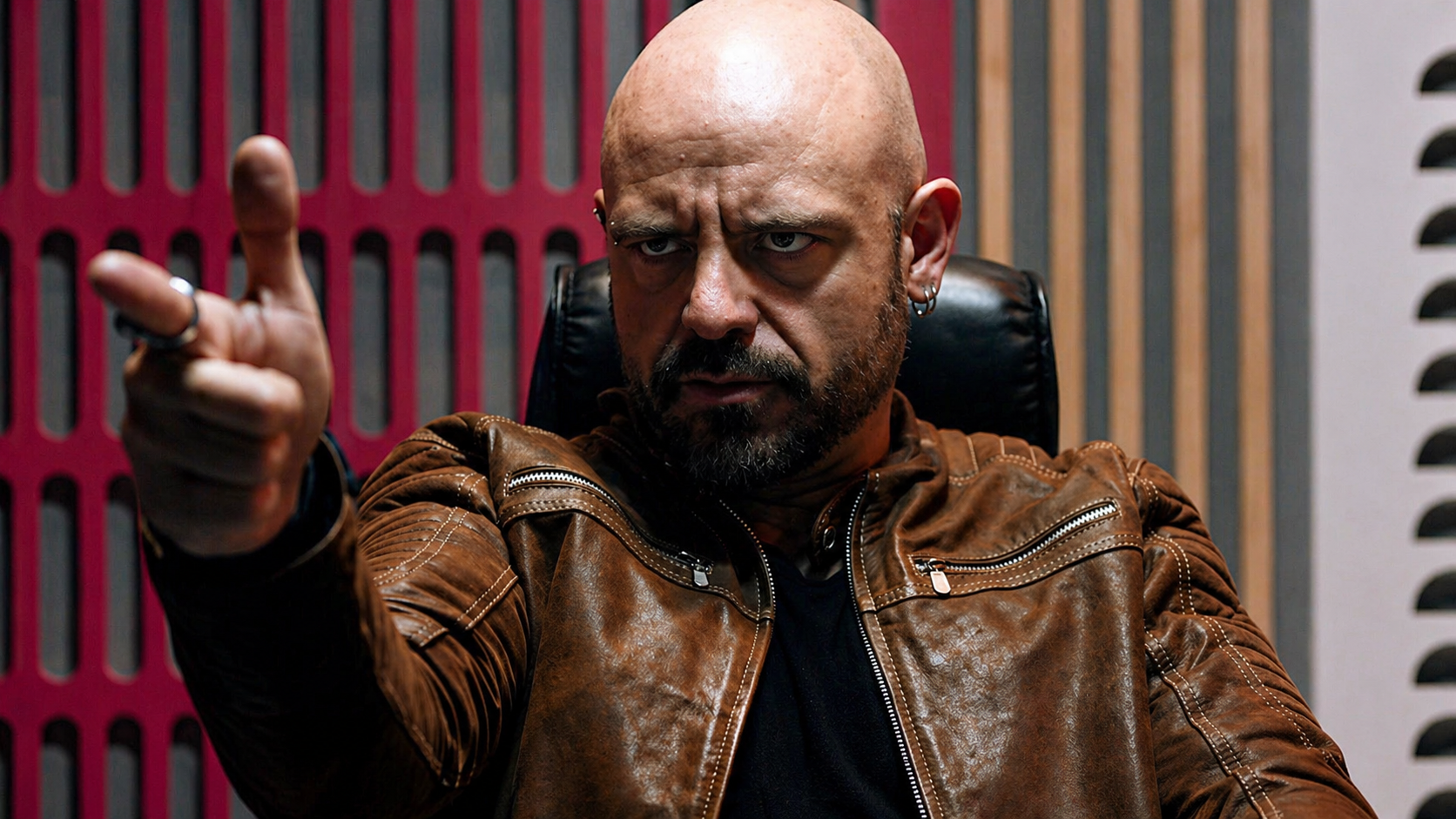 Hombre calvo con barba, vistiendo chaqueta de cuero marrón, sentado en una oficina, apuntando hacia adelante con expresión intensa.