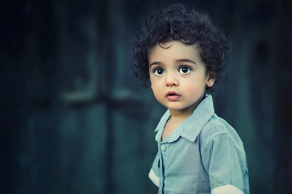 Niño de cabello rizado y expresión asombrada, vestido con camiseta de manga larga a rayas, en un fondo oscuro y despejado.