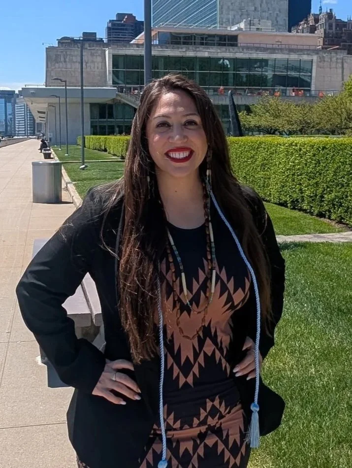 A woman smiling outdoors in front of a modern cityscape with tall buildings and greenery, wearing a black blazer and patterned dress.