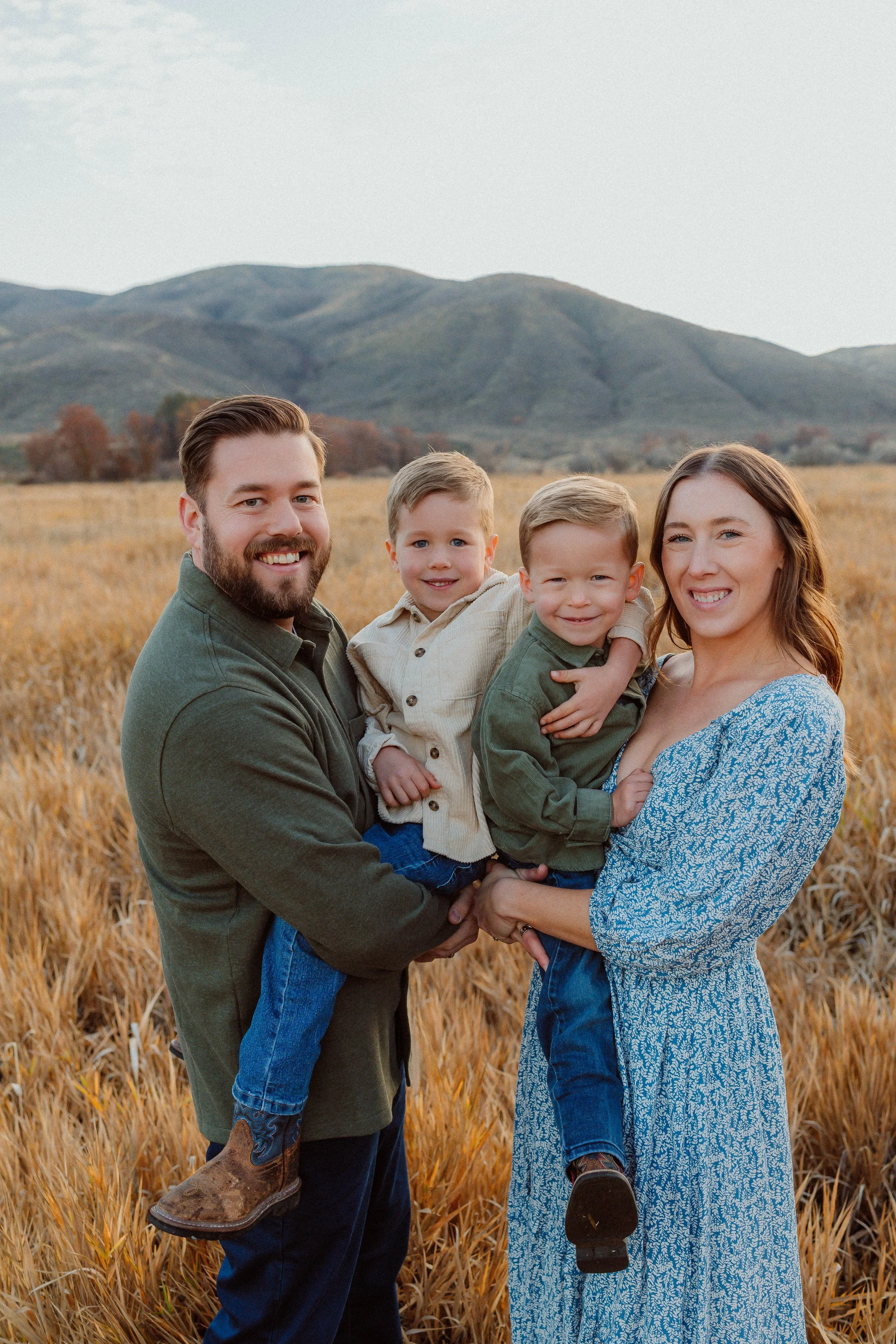 A smiling family of four, including a man, a woman, and two young boys, standing in a golden wheat field with mountains in the background during daytime.