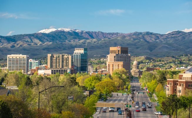 Downtown cityscape with buildings, trees, and mountains in the background on a clear day.