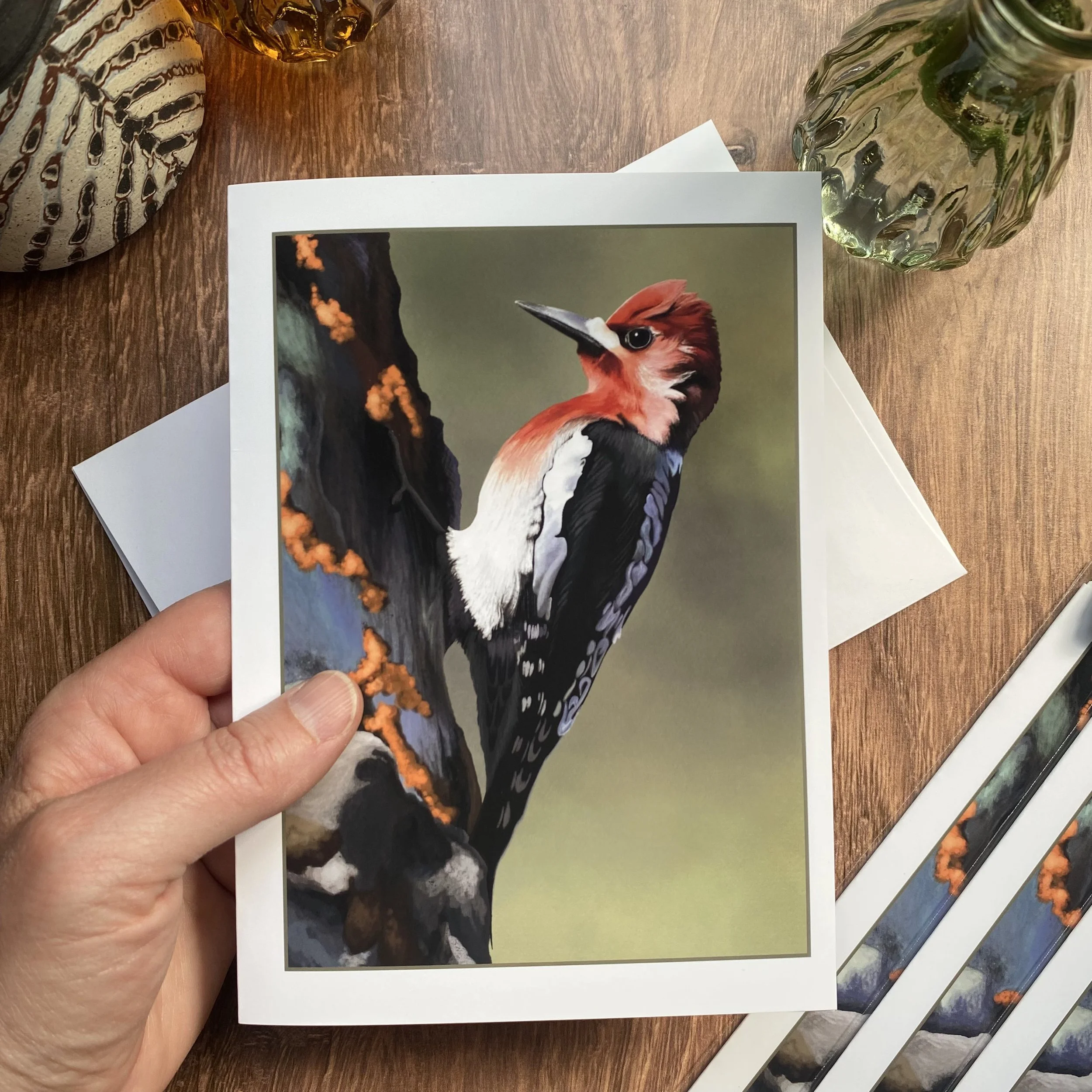 A photograph of a printed picture of a bird, specifically a woodpecker, held by a hand over a wooden surface. The picture highlights the bird with vibrant red and black feathers and a white patch on its cheek and chest.