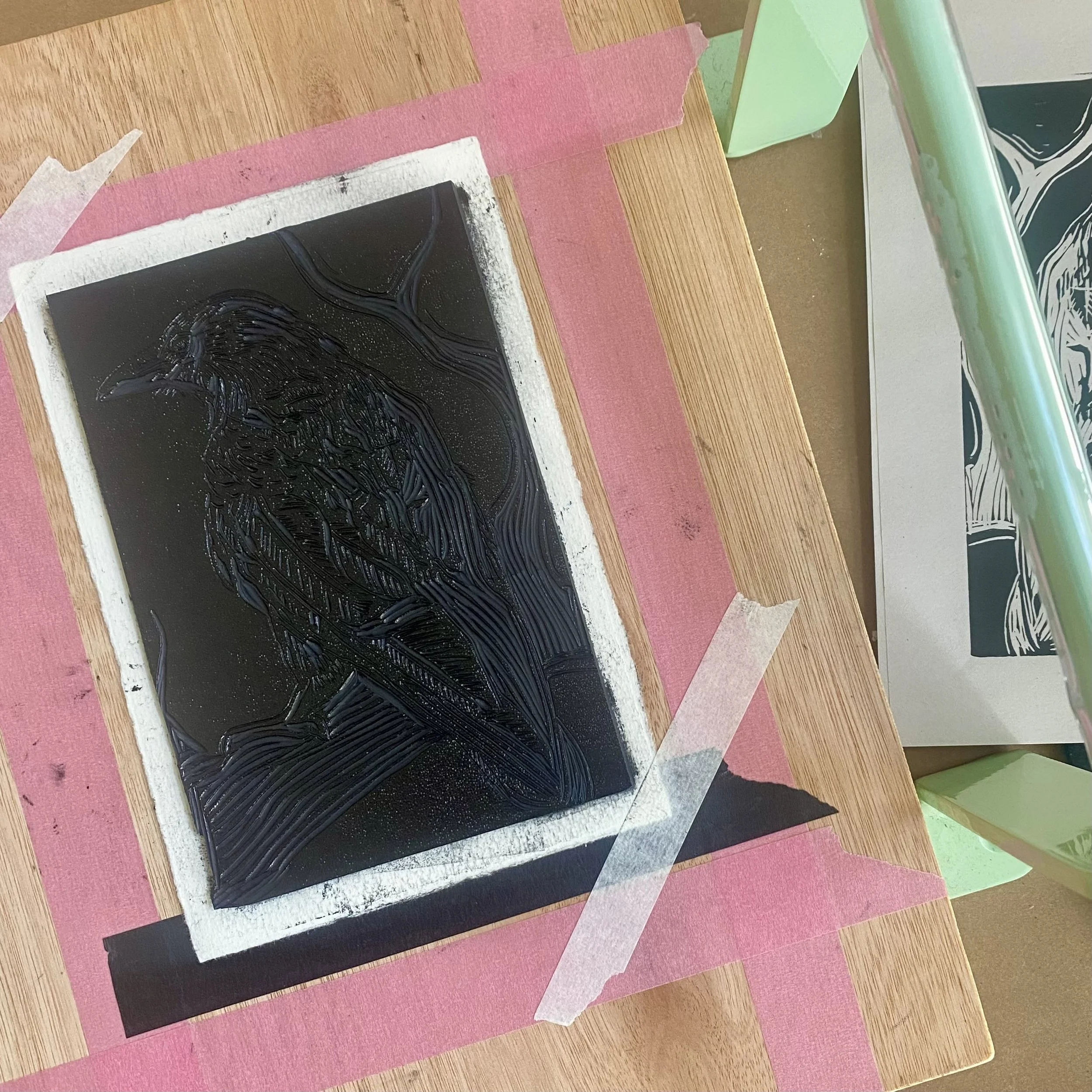 An art print with black and white abstract designs placed on a wooden table with pink painter's tape around it and green furniture nearby.