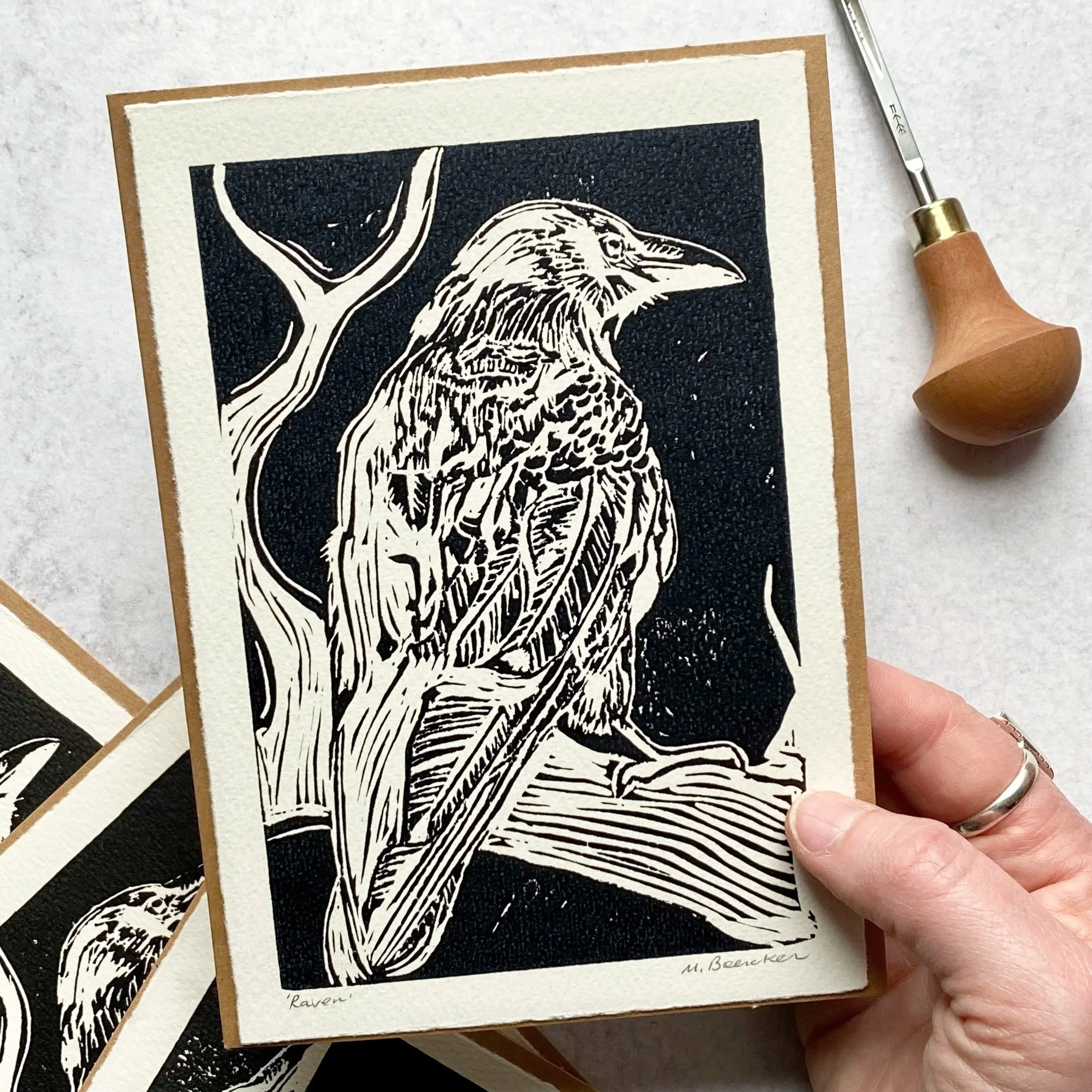 A hand holding two small prints of a black and white illustration of a raven, with one print partially overlapping the other, on a light-colored surface.