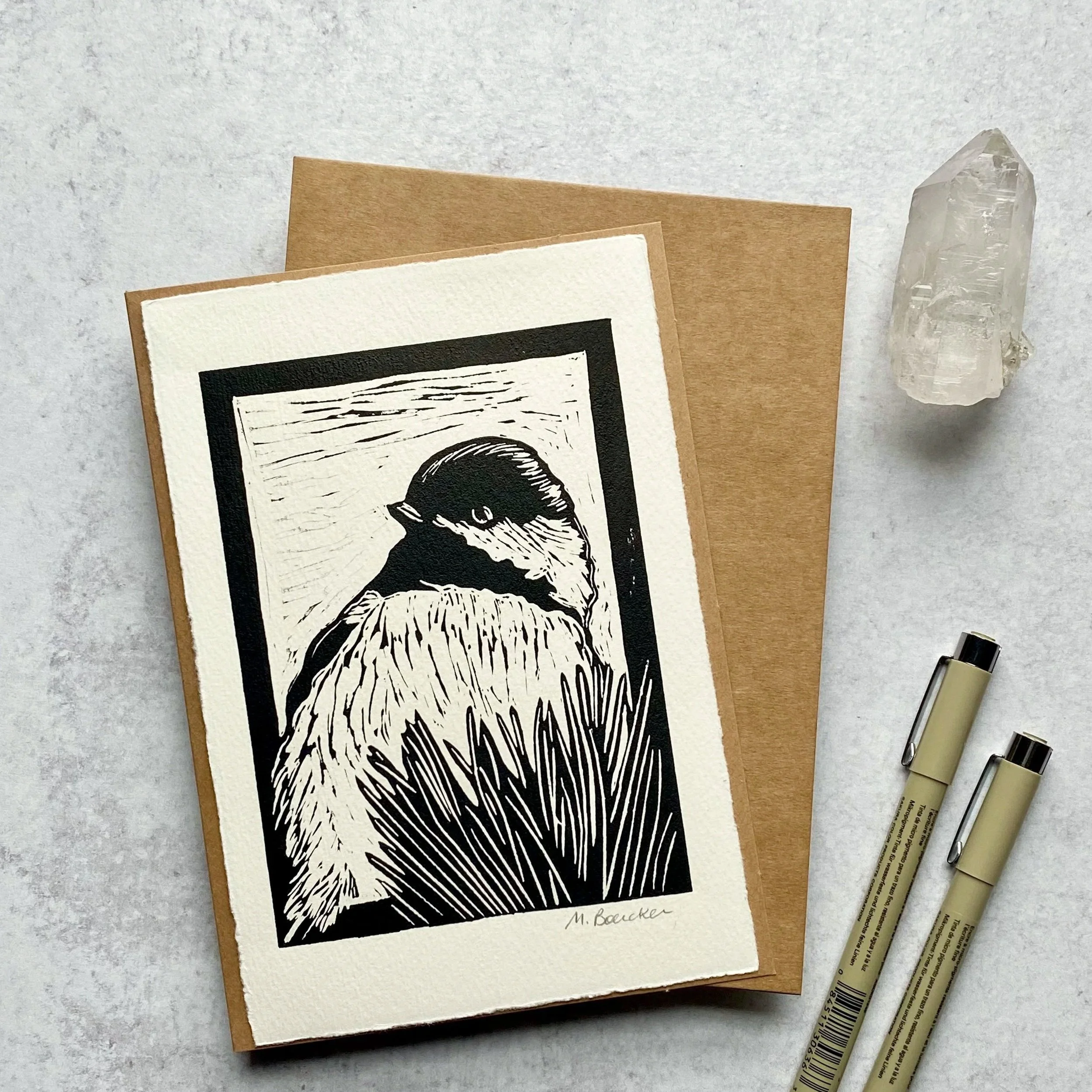 A set of three beige and white textured stationery cards with a black and white landscape illustration and handwritten signature, along with three metallic pens and a clear quartz crystal on a gray surface.