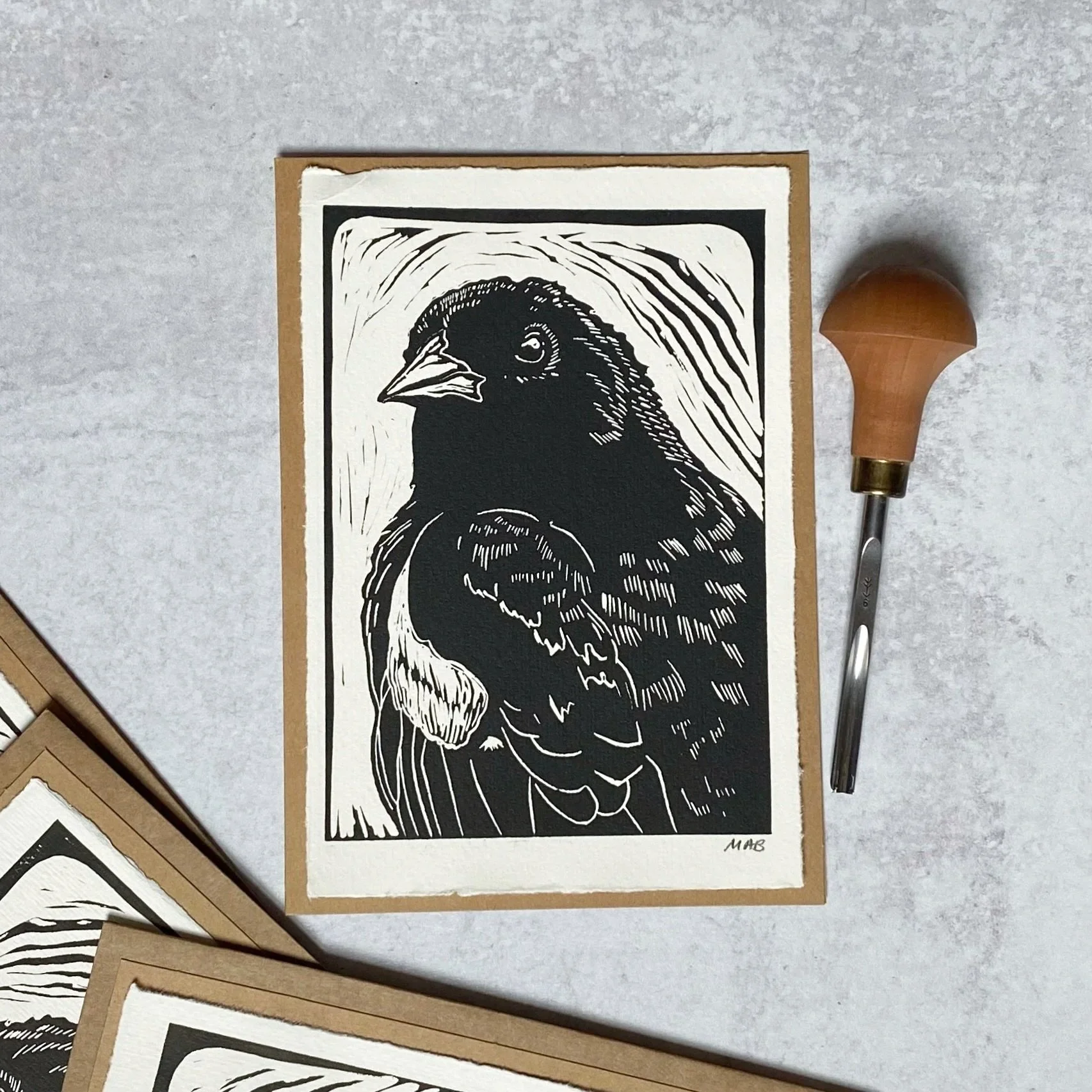 A black and white linocut print of a bird, likely a sparrow, with a beak pointing to the left, in a frame with a textured background. There is a wooden-handled carving tool next to the print.