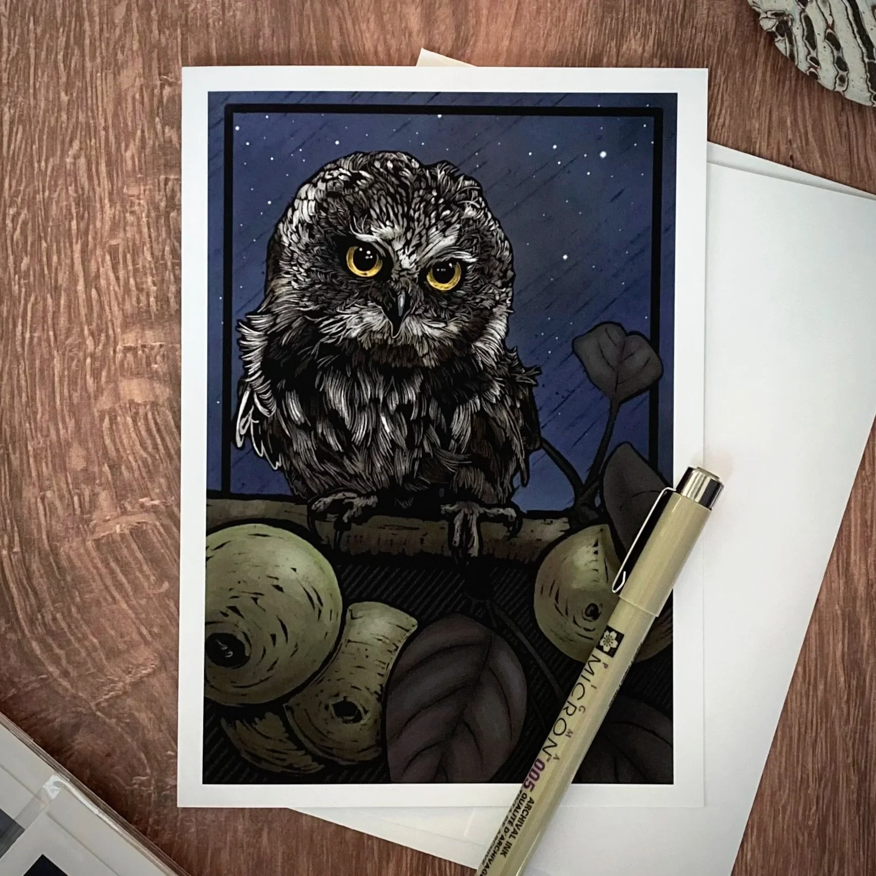Illustration of an owl with a dark blue night sky background, stars, and a branch. The owl has detailed feathers and large, yellow eyes. There are some objects including a white paper, a fur-covered item, and a beige marker pen around the illustratio
