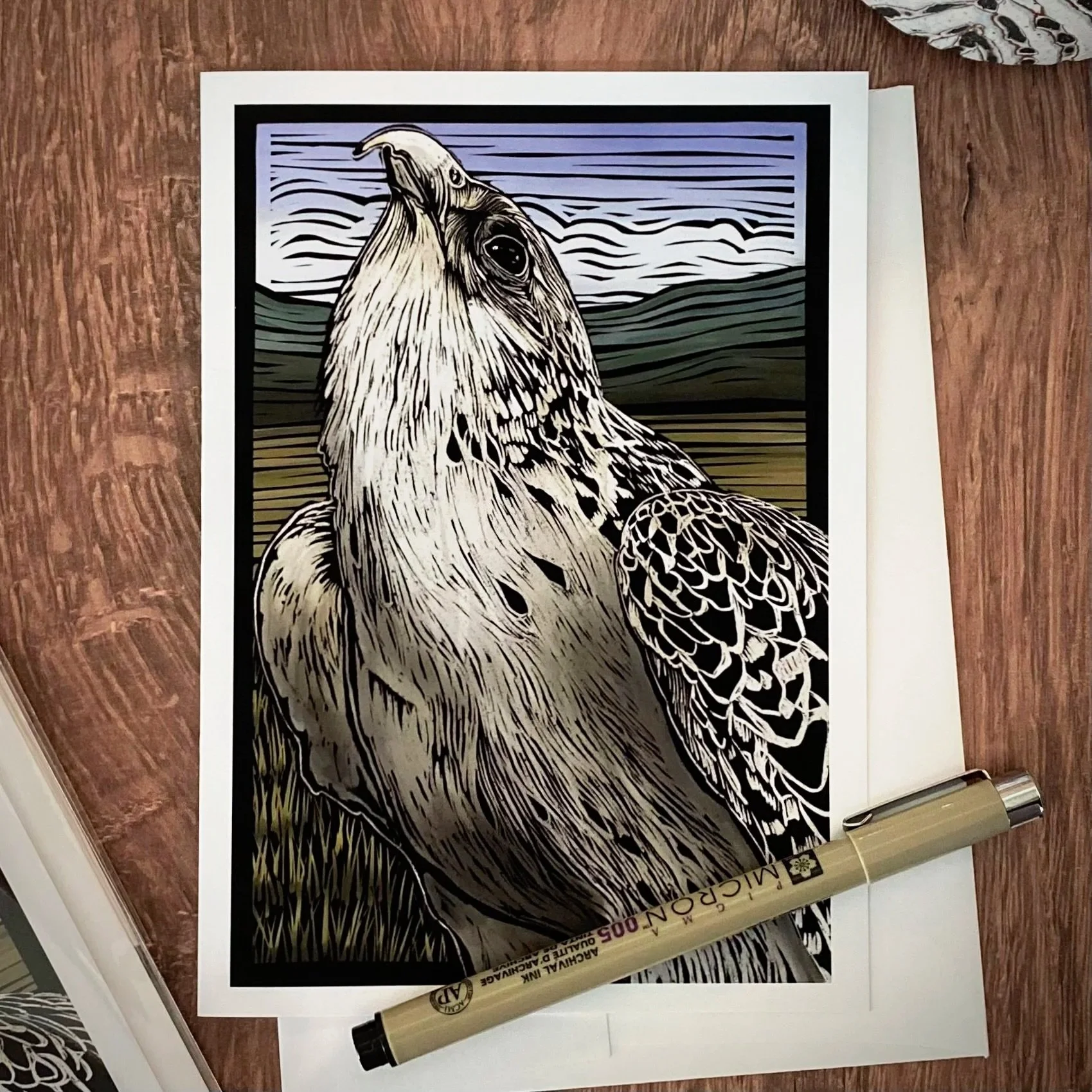 A black and white illustration of a bird, possibly a hawk or eagle, with detailed feathers, set against a landscape background with lines representing the sky and mountains. A metallic marker pen lies at the bottom of the illustration.