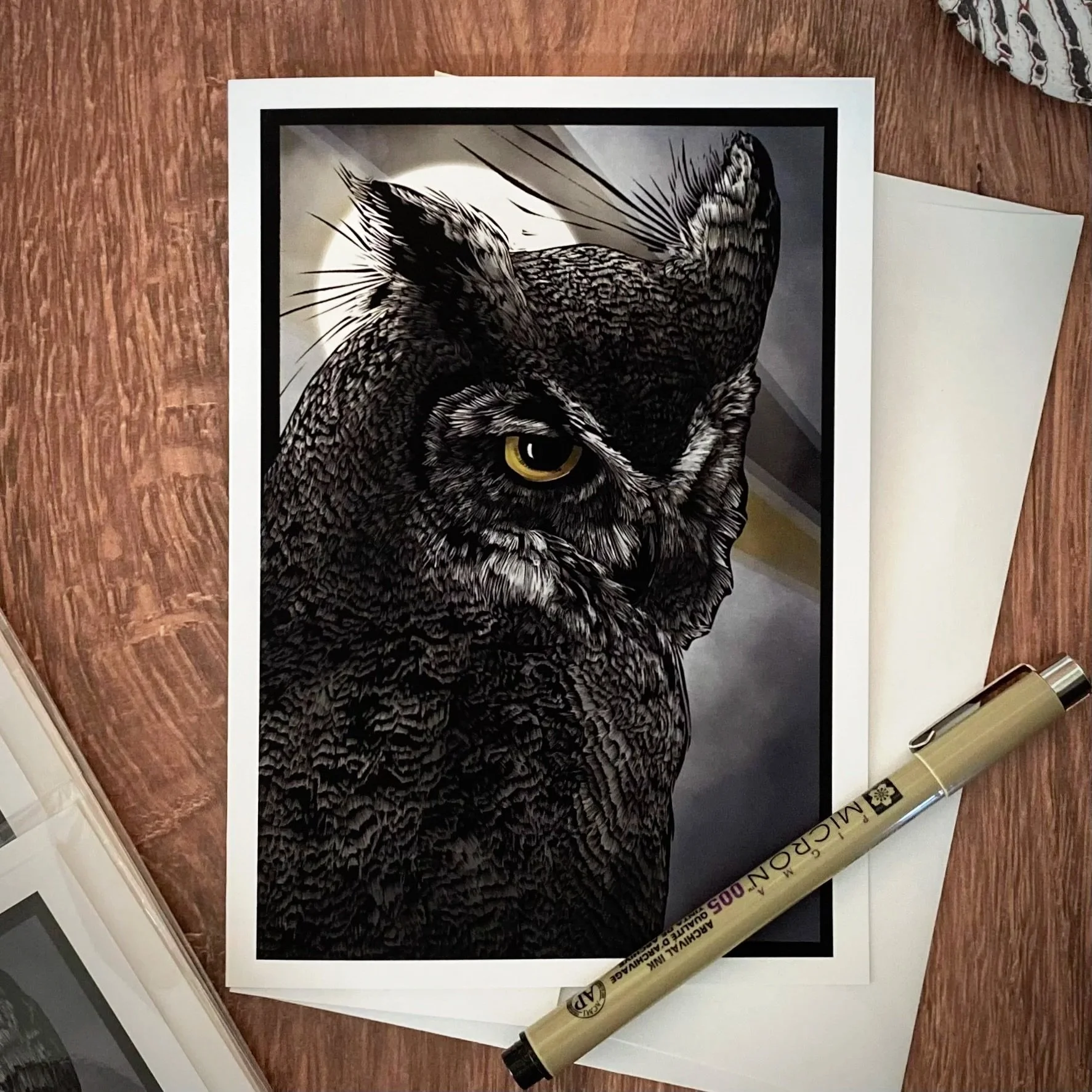 A printed black and white photo of an owl with yellow eyes, laid on a wooden surface alongside a tan marker and some papers.