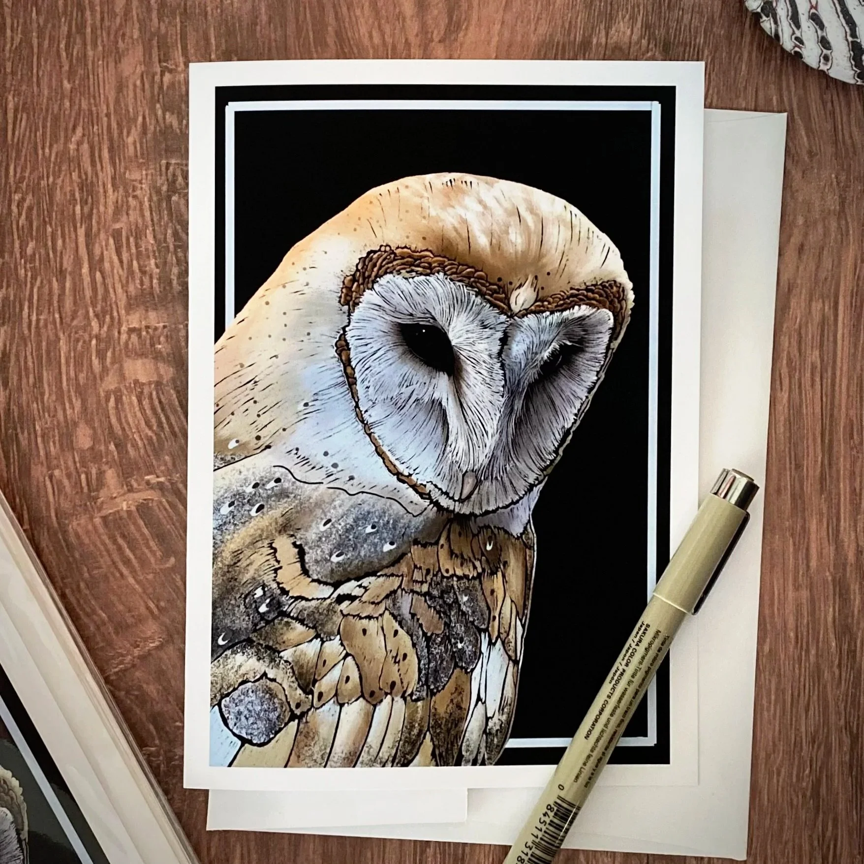 A detailed illustration of a barn owl's face, wing, and a portion of its body, on glossy paper next to a marker pen on a wooden surface.