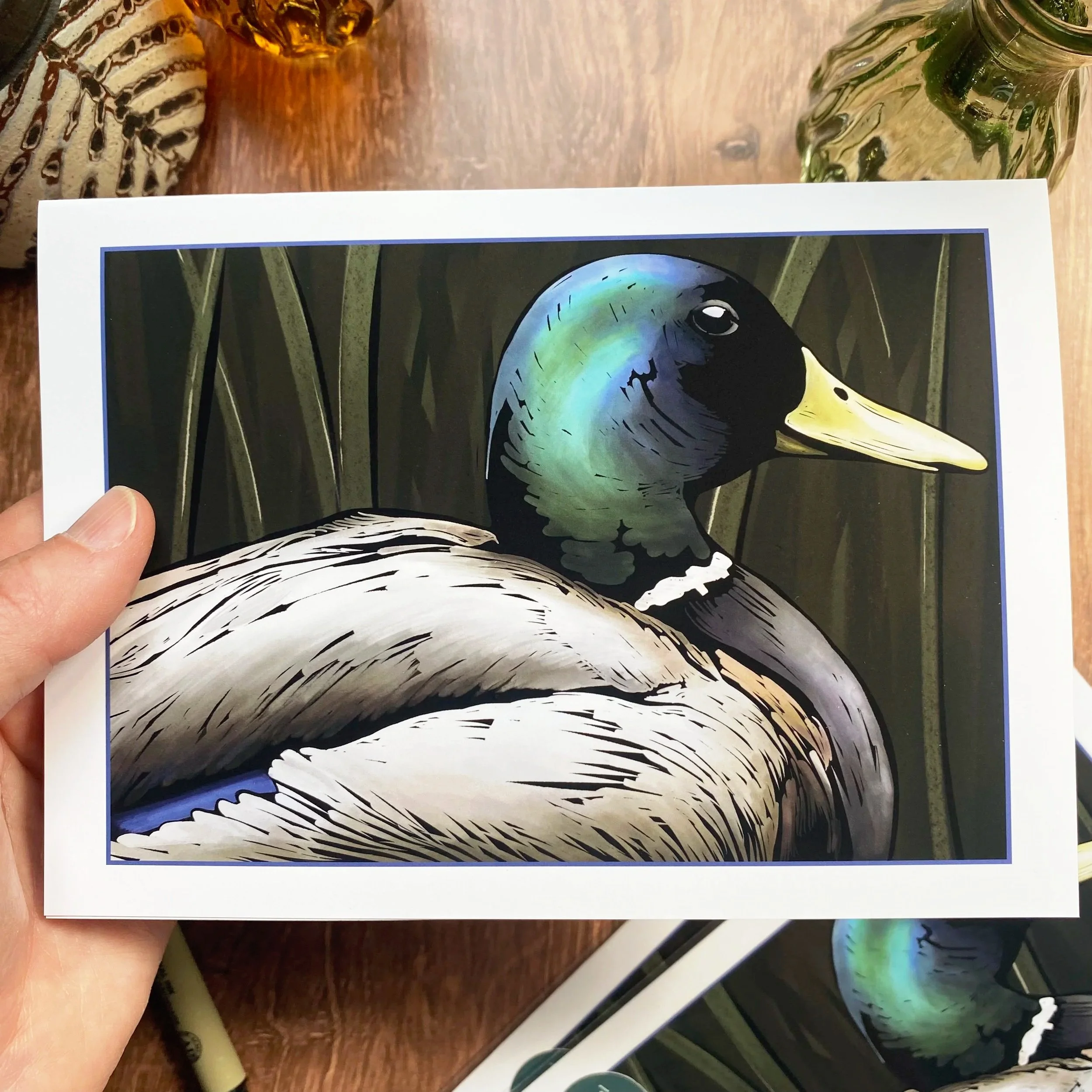A printed poster of a colorful illustration of a bird, specifically a duck, with a green head, yellow beak, and black neck, placed on a wooden surface with glass objects nearby.