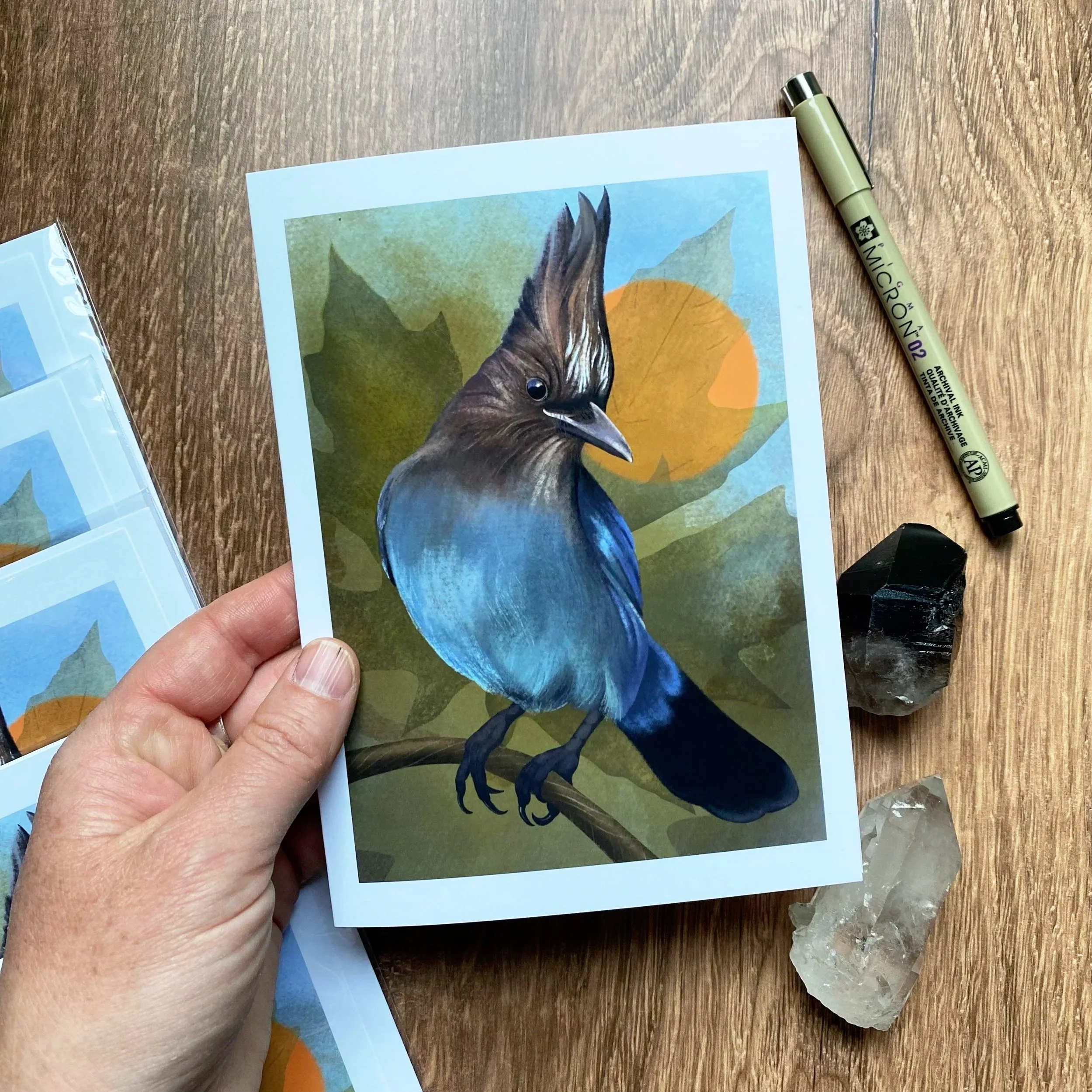 Close-up of a printed illustration of a bird on a piece of paper, with a hand holding the paper, and a beige marker pen laying on a wooden table alongside some rocks.