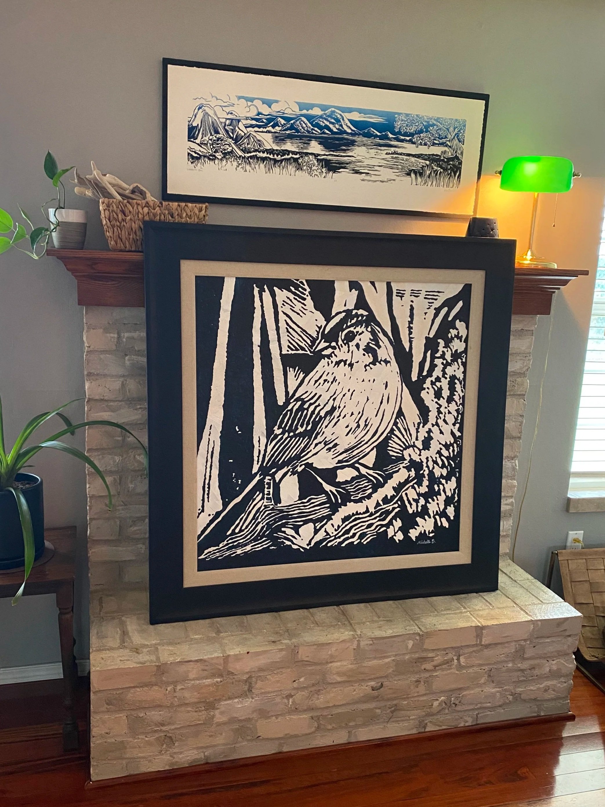 A framed black-and-white line drawing of a bird sitting on a branch, leaning against a brick fireplace, with a decorative lamp and plants nearby.