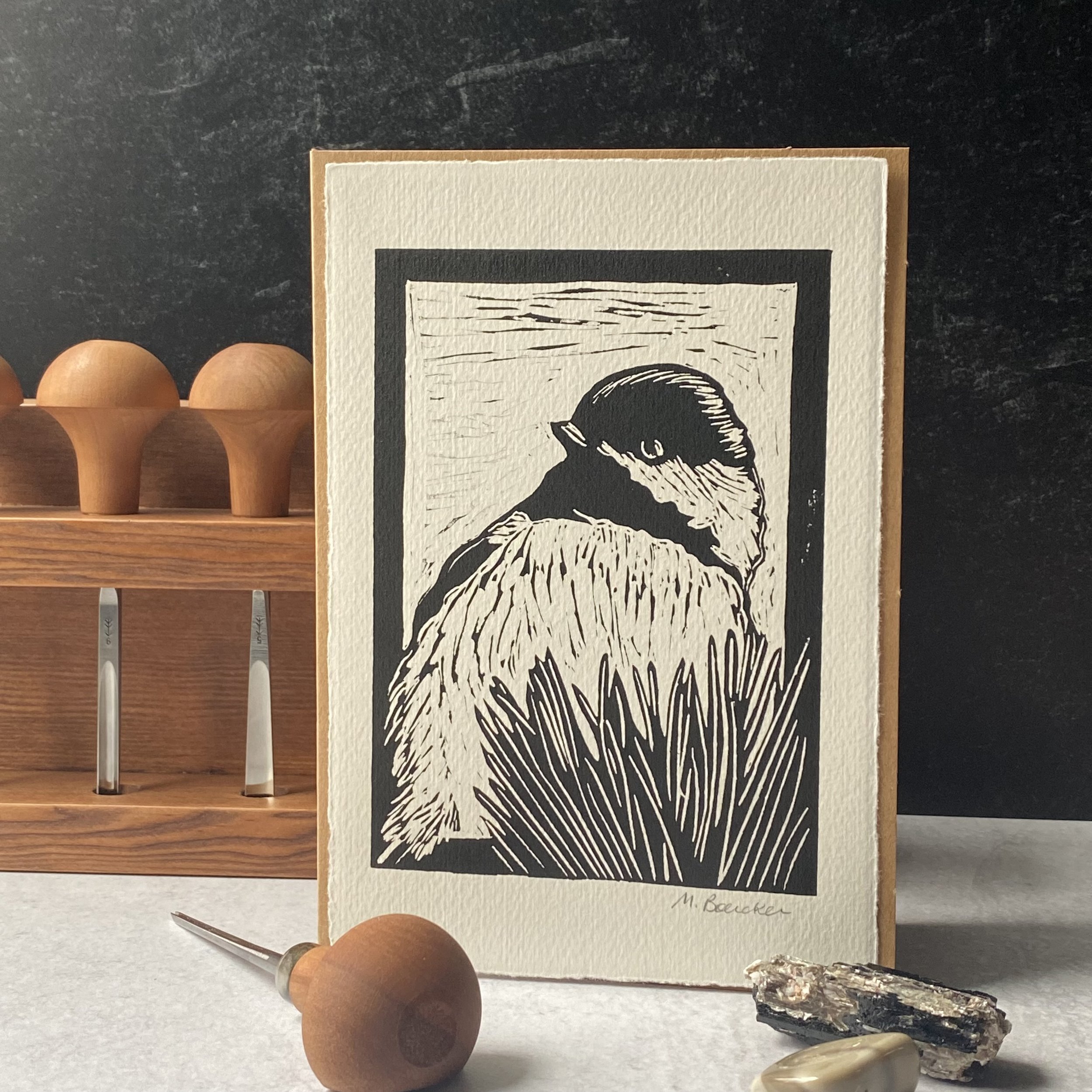 A black and white illustration of a bearded man's profile, on a card, with wooden objects and stones placed nearby.