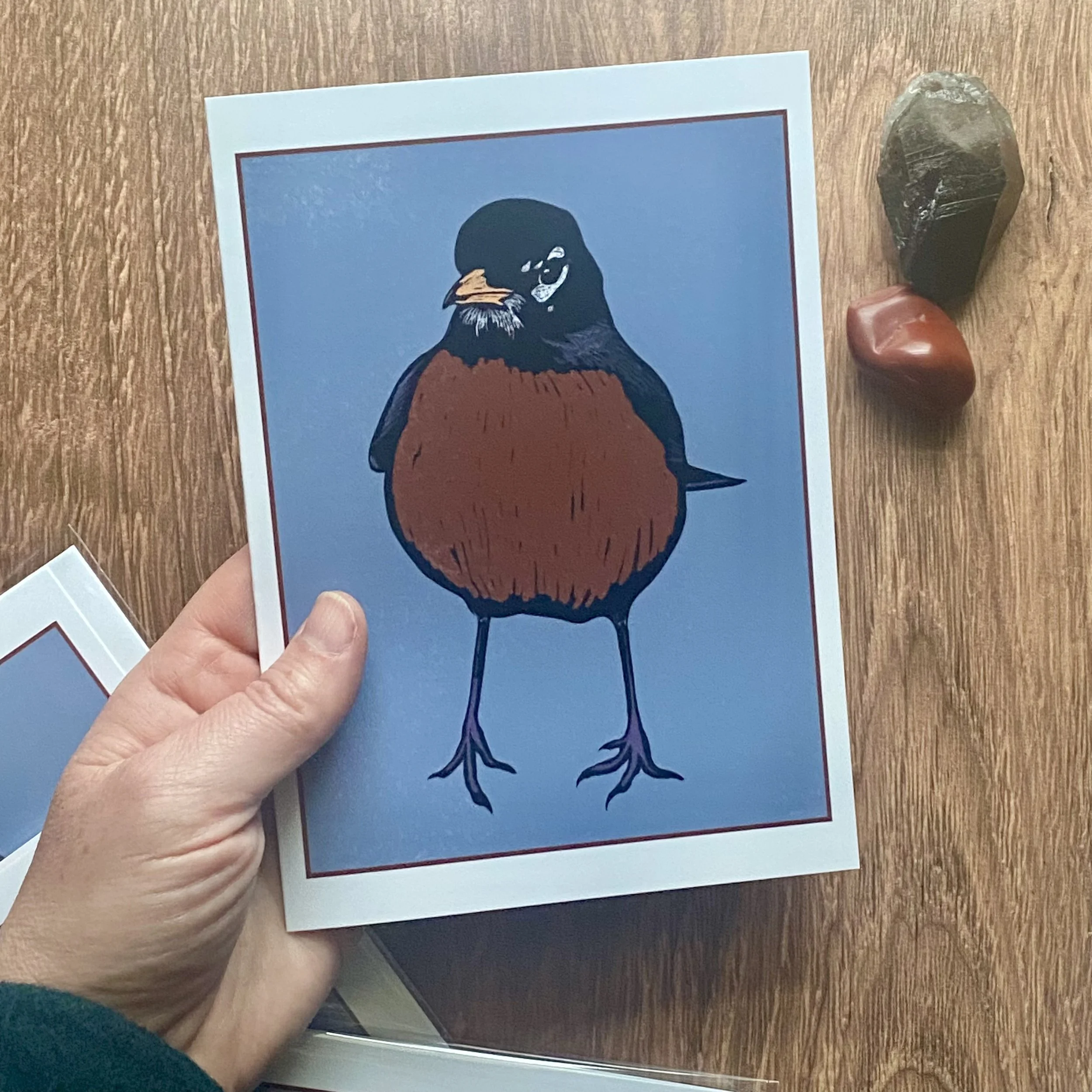 A hand holding a postcard with an illustration of a bird against a blue background, placed on a wooden surface with three crystals nearby.