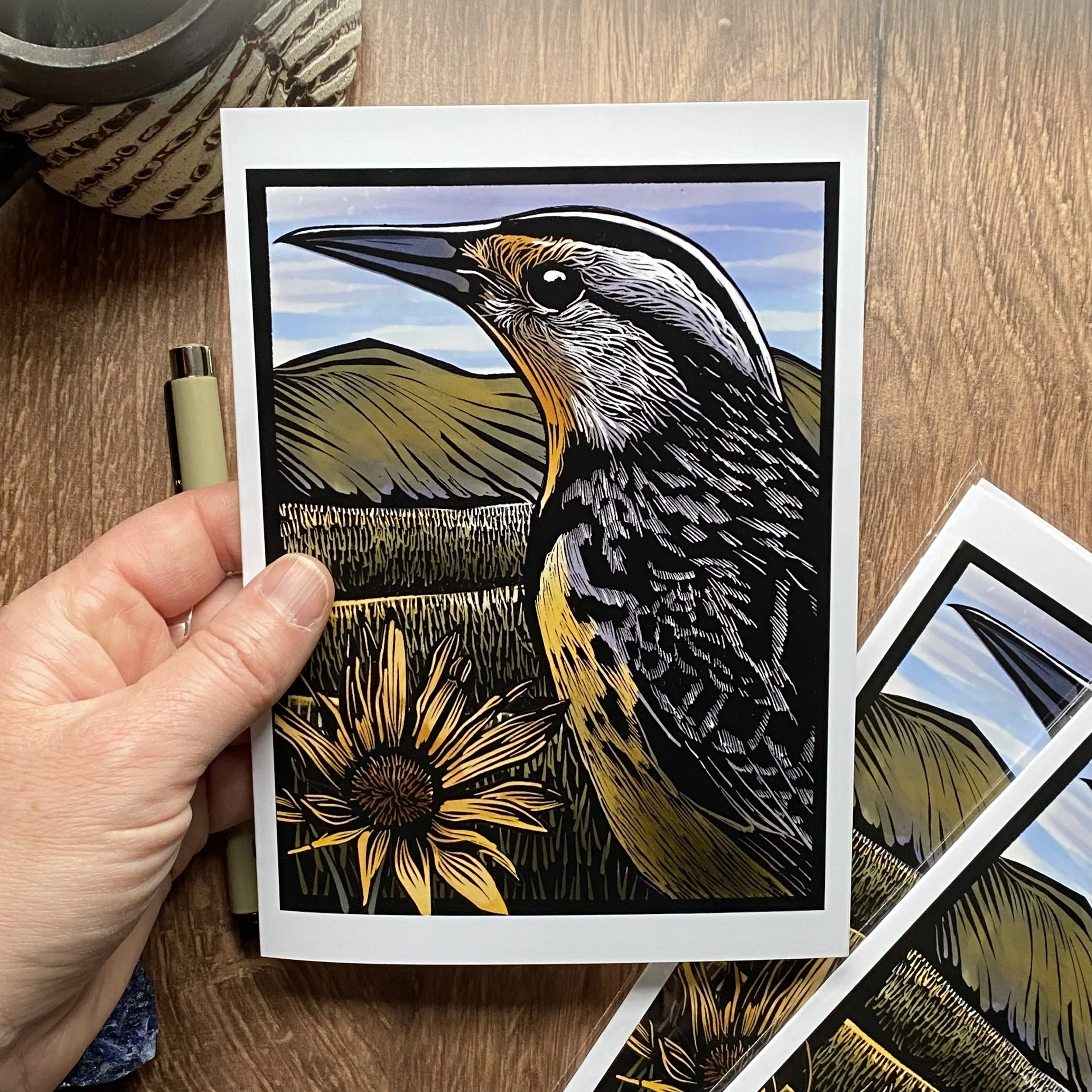 Close-up of a person holding a print of a stylized bird illustration, with additional prints and art supplies on a wooden surface.