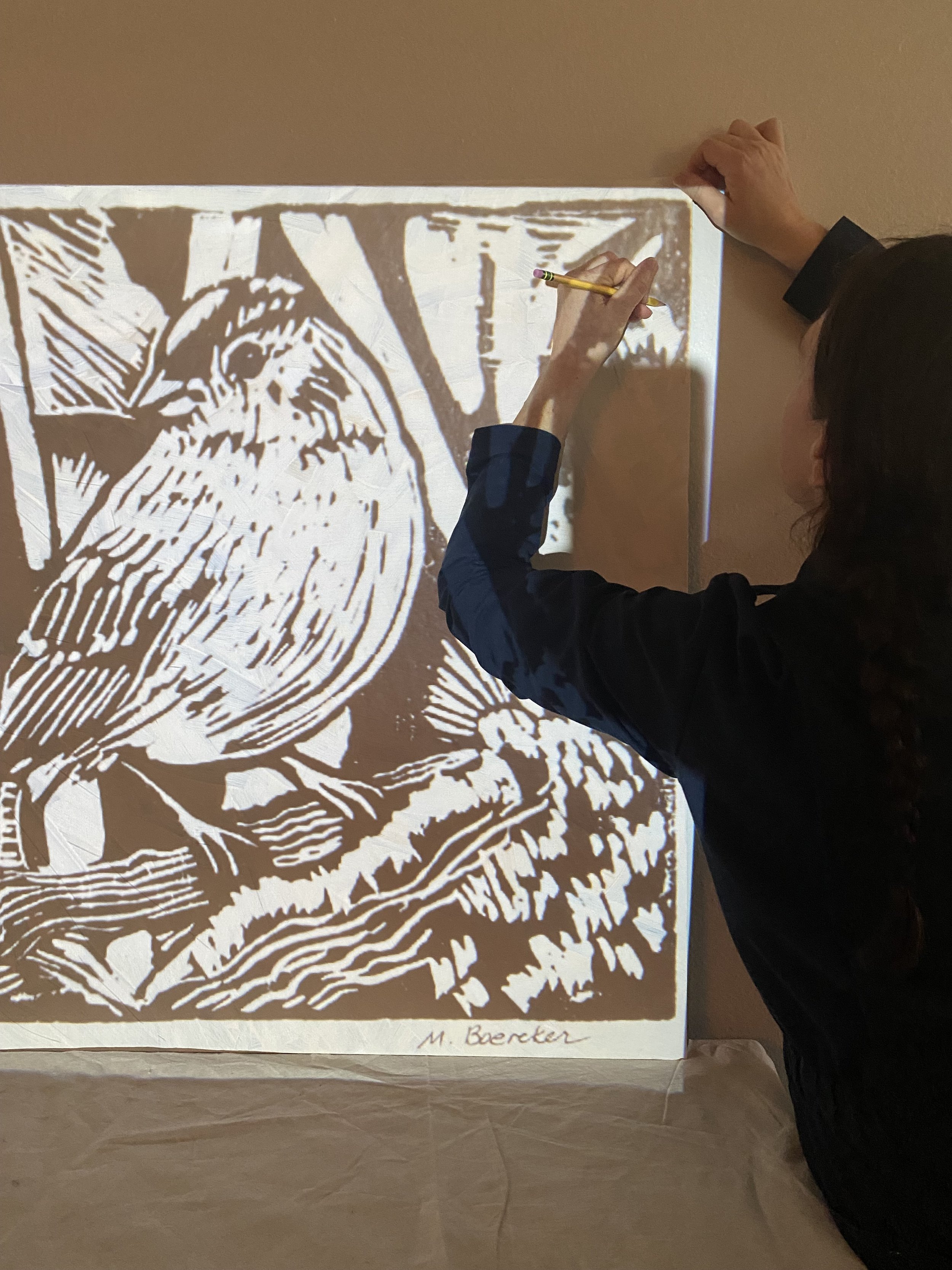 A person using a yellow pencil to trace a black and white print of a lion's face and mane on a beige wall. The print appears to be a woodcut or linocut style with bold lines. The print has the signature 'M. Becker' at the bottom.