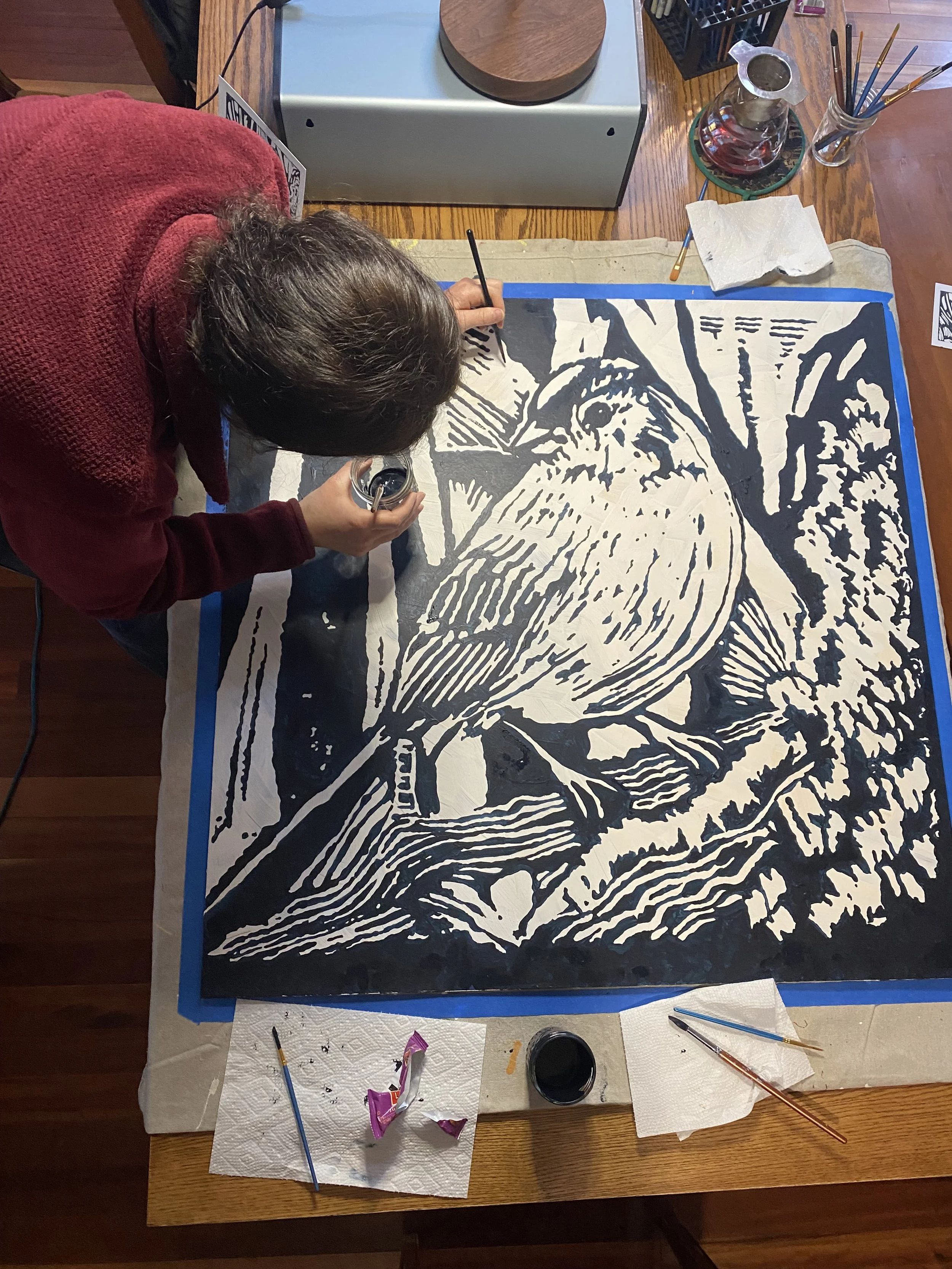 A person in a red sweater is working on a large black and white painting of a dog, possibly a Labrador retriever, on a wooden table with art supplies such as brushes, paint, and paper towels.