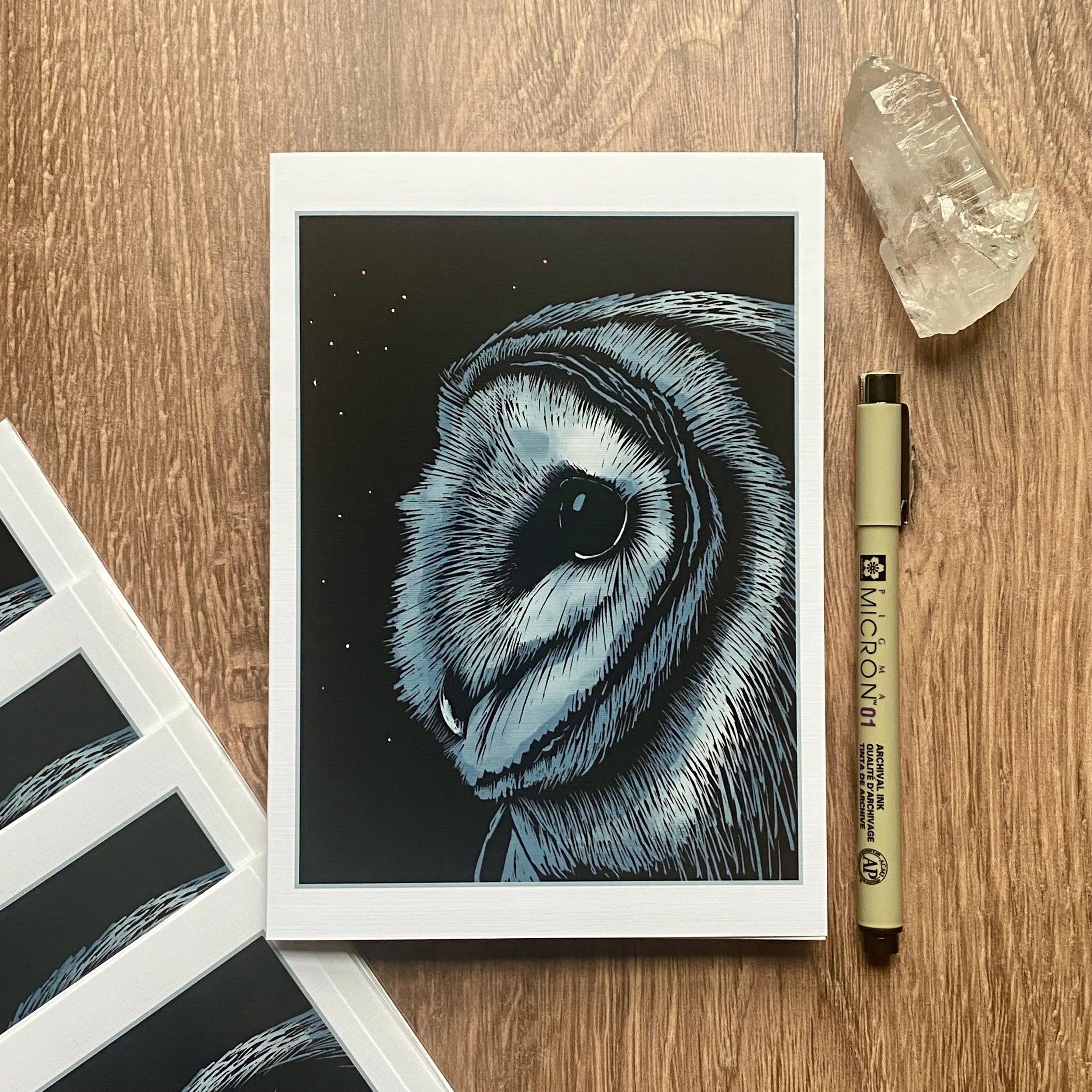 A print of a black and white drawing of a koala bear's face on a black background. The print is framed with a white border. To the right of the print, there is a clear quartz crystal and a beige marker with black and purple text on it, placed on a wo