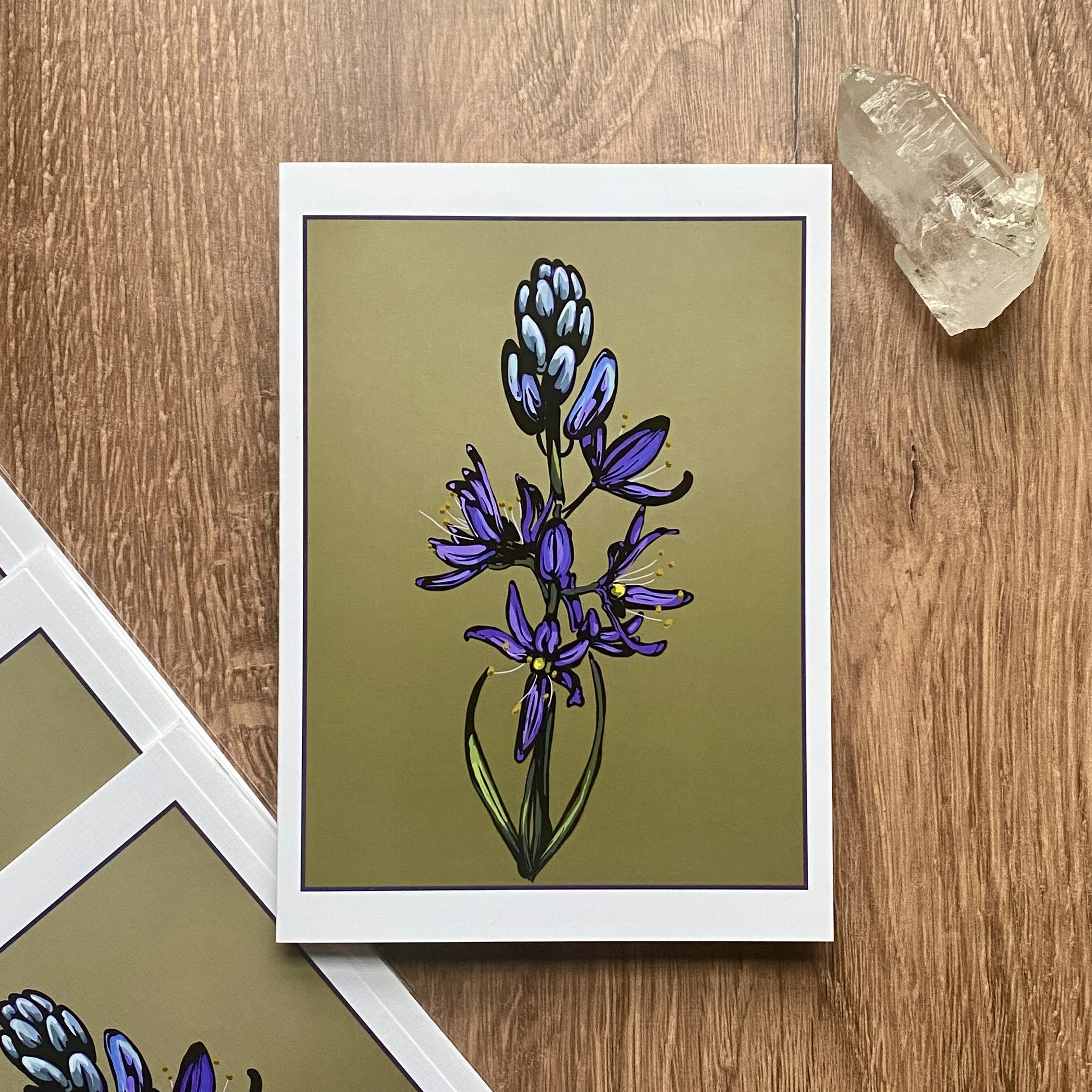 A printed botanical illustration of a purple lavender flower on a pale green background, placed on a wooden surface with an additional clear quartz crystal and loose paper nearby.