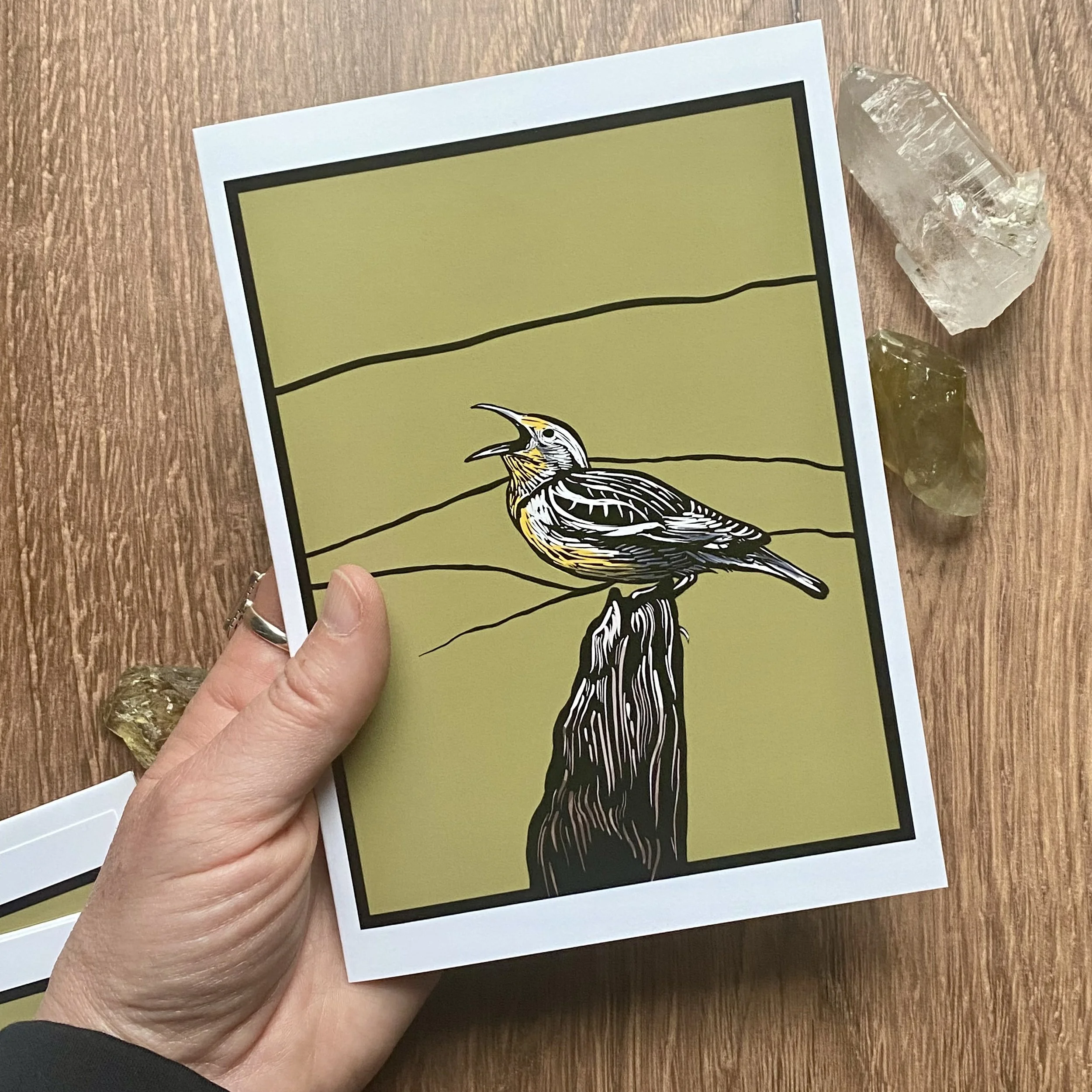 Close-up of a hand holding a printed card of a bird illustration with a yellow background, with three uncut gemstones on a wooden surface.