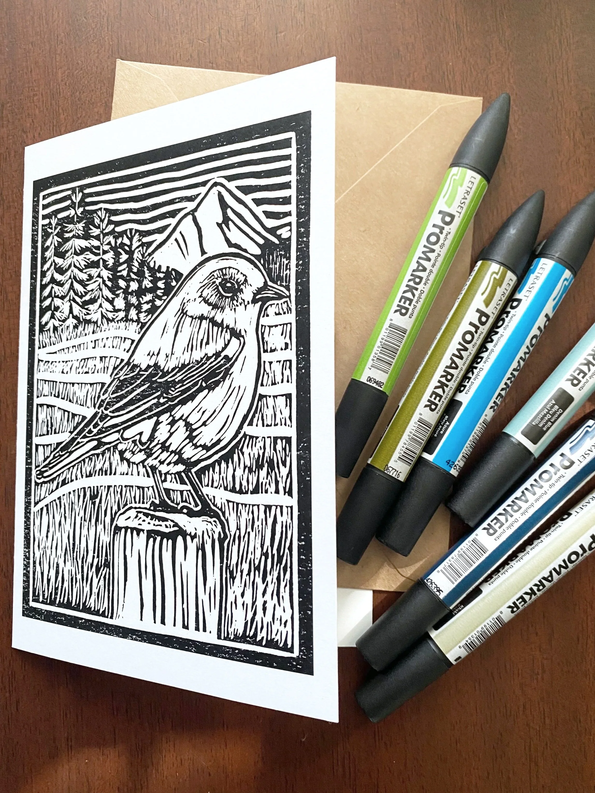 A black and white linocut print of a bird, likely a mountain bird, with a mountain and trees in the background, on a piece of white paper. Four colorful markers are laid out next to the paper on a wooden surface.
