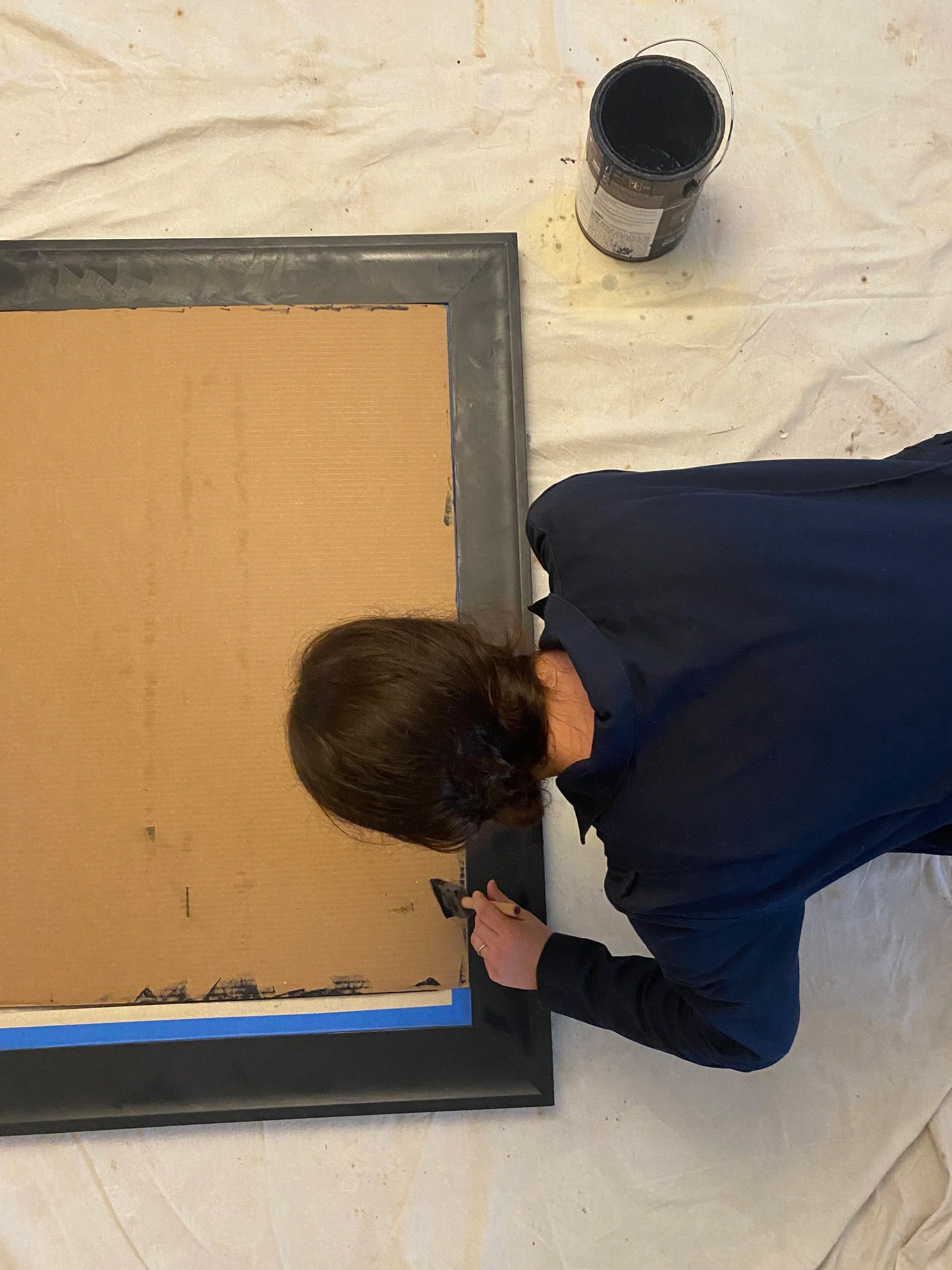 A person with short brown hair and a navy blue jacket is kneeling on the floor and taking a picture of a framed artwork on the wall with a smartphone.