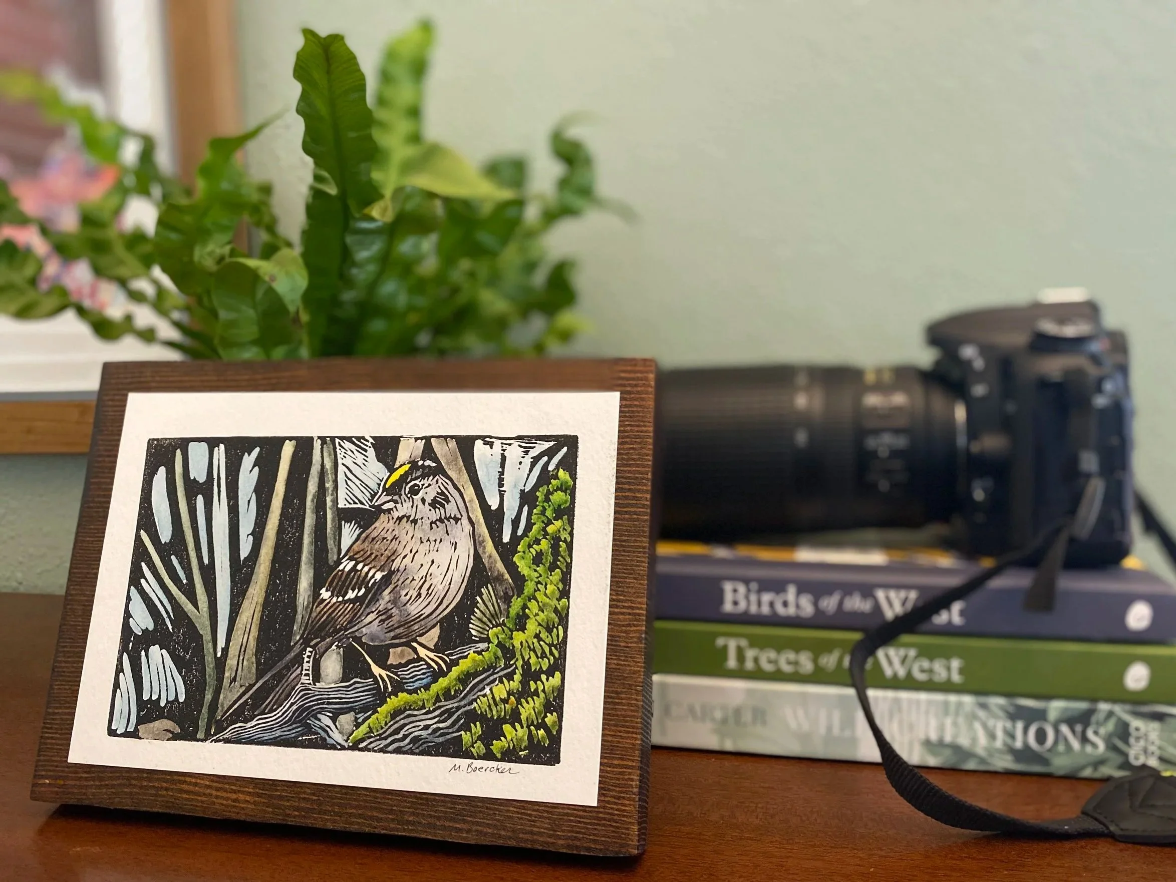A small illustration of a bird sitting on a branch, surrounded by trees, with green foliage nearby, displayed on a wooden surface.