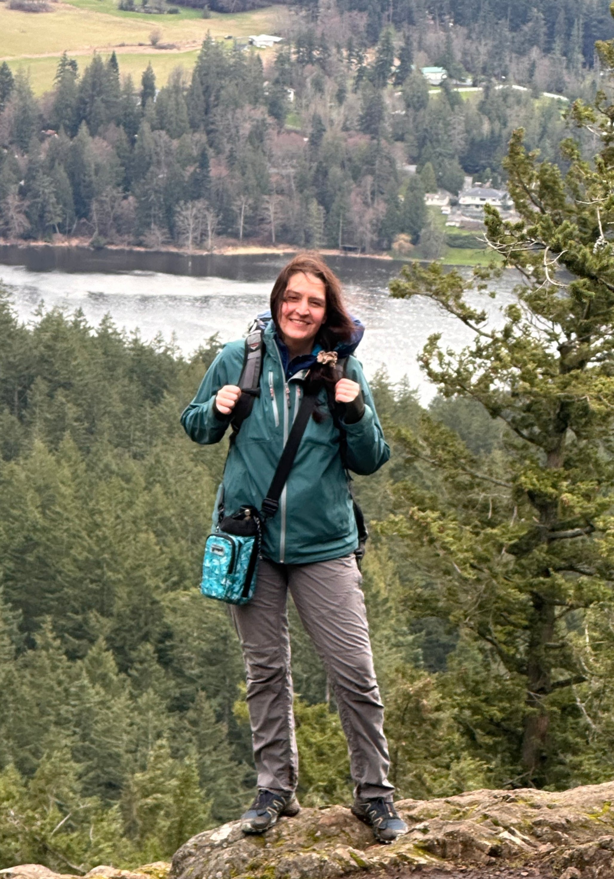A woman standing on a rock, smiling, with a backpack, in a forested outdoor setting near a river.