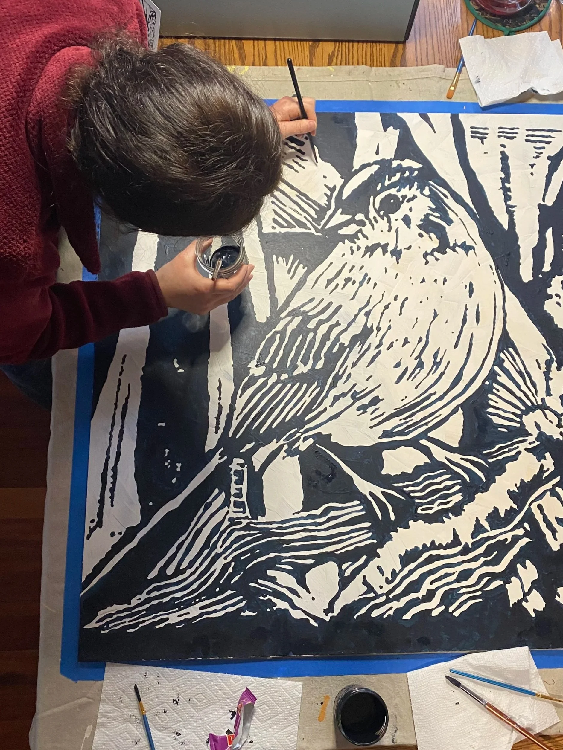 An artist creating a woodcut print of a lion's face, using black ink on paper, surrounded by painting tools and paper towels.