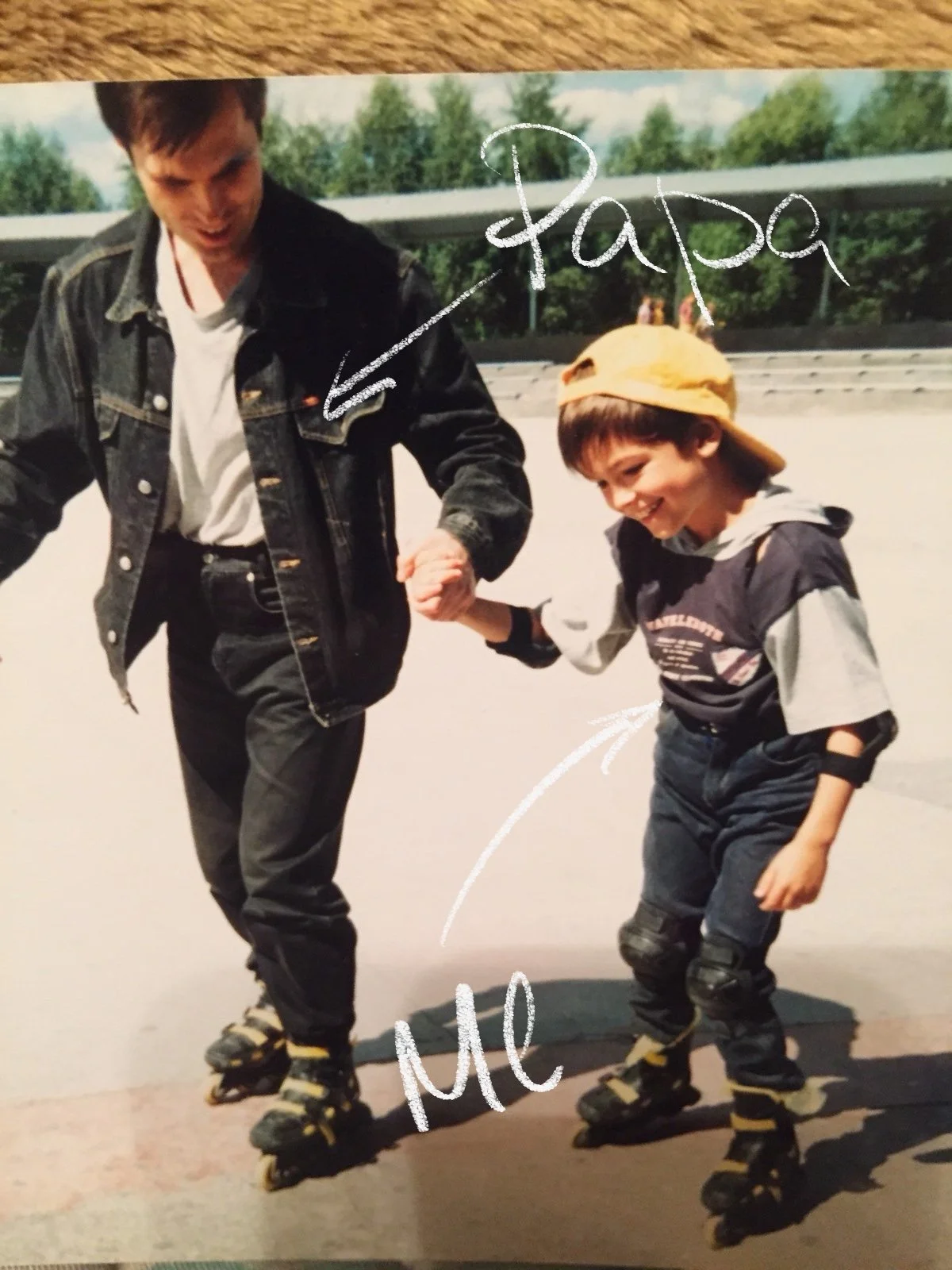 A young boy and an older male skating together outdoors, holding hands and smiling. The boy is wearing a yellow cap and casual clothing, and both are on rollerblades. The background includes trees and a railing.