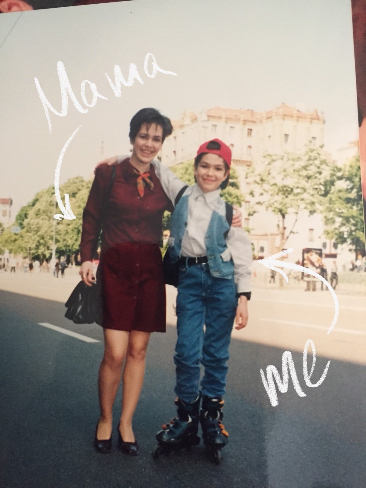 A woman and a boy standing on a city street, smiling. The woman has short dark hair, wears a burgundy outfit, and carries a black purse. The boy is wearing rollerblades, a white shirt, a denim vest, a red backwards cap, and has a backpack. There are trees and a historic building in the background. The photo has handwritten text saying "Mama" and "me" with arrows pointing to the woman and the boy, respectively.