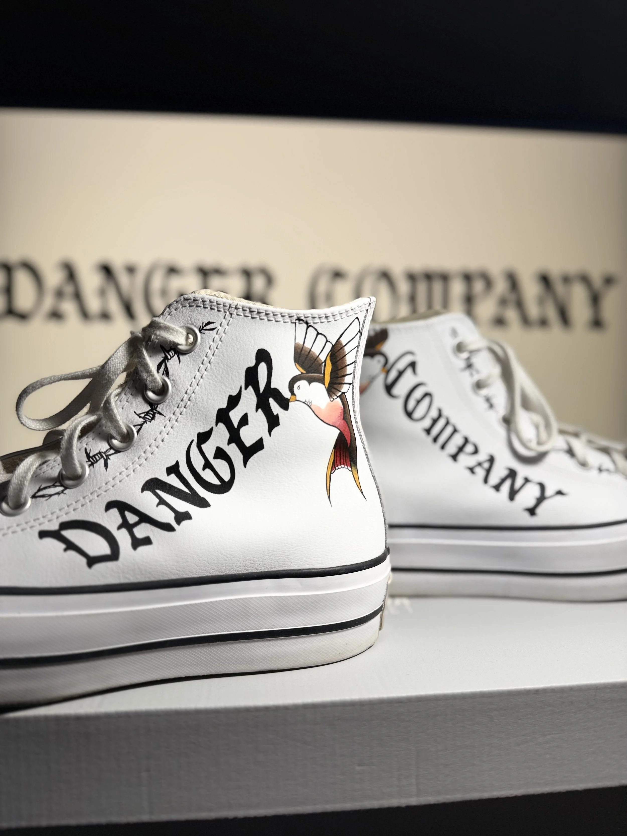 Close-up of white high-top sneakers with black text and a colored bird illustration, placed on a shelf against a black and beige background.