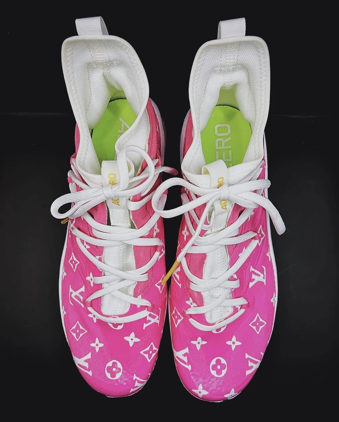 Pair of pink Louis Vuitton designer sneakers with white laces and logo pattern, on a black background.