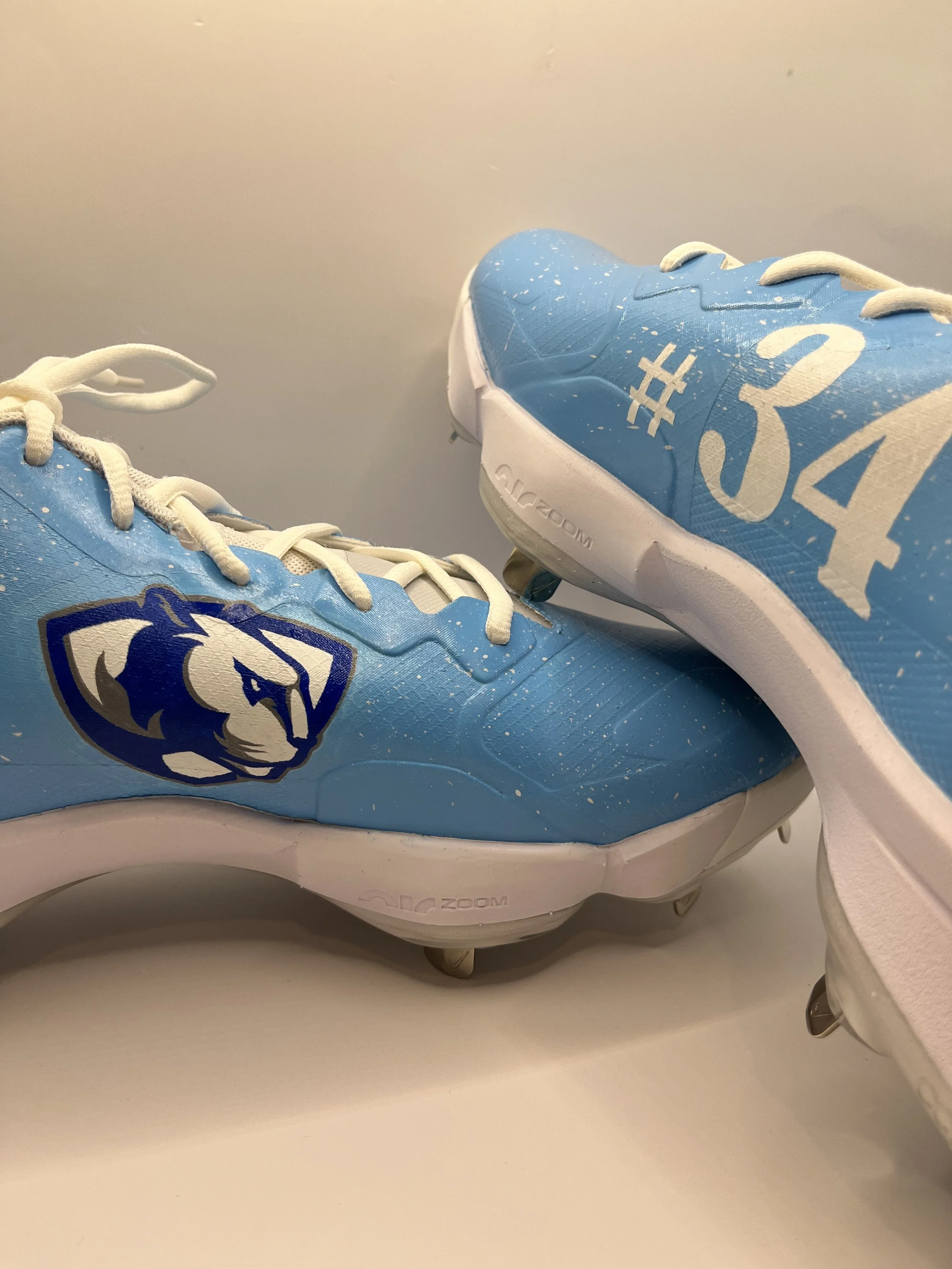 A pair of blue athletic shoes with white laces and a lion logo on the side. One shoe has white text with the hashtag #34 on the side. The shoes are sitting on a white surface.