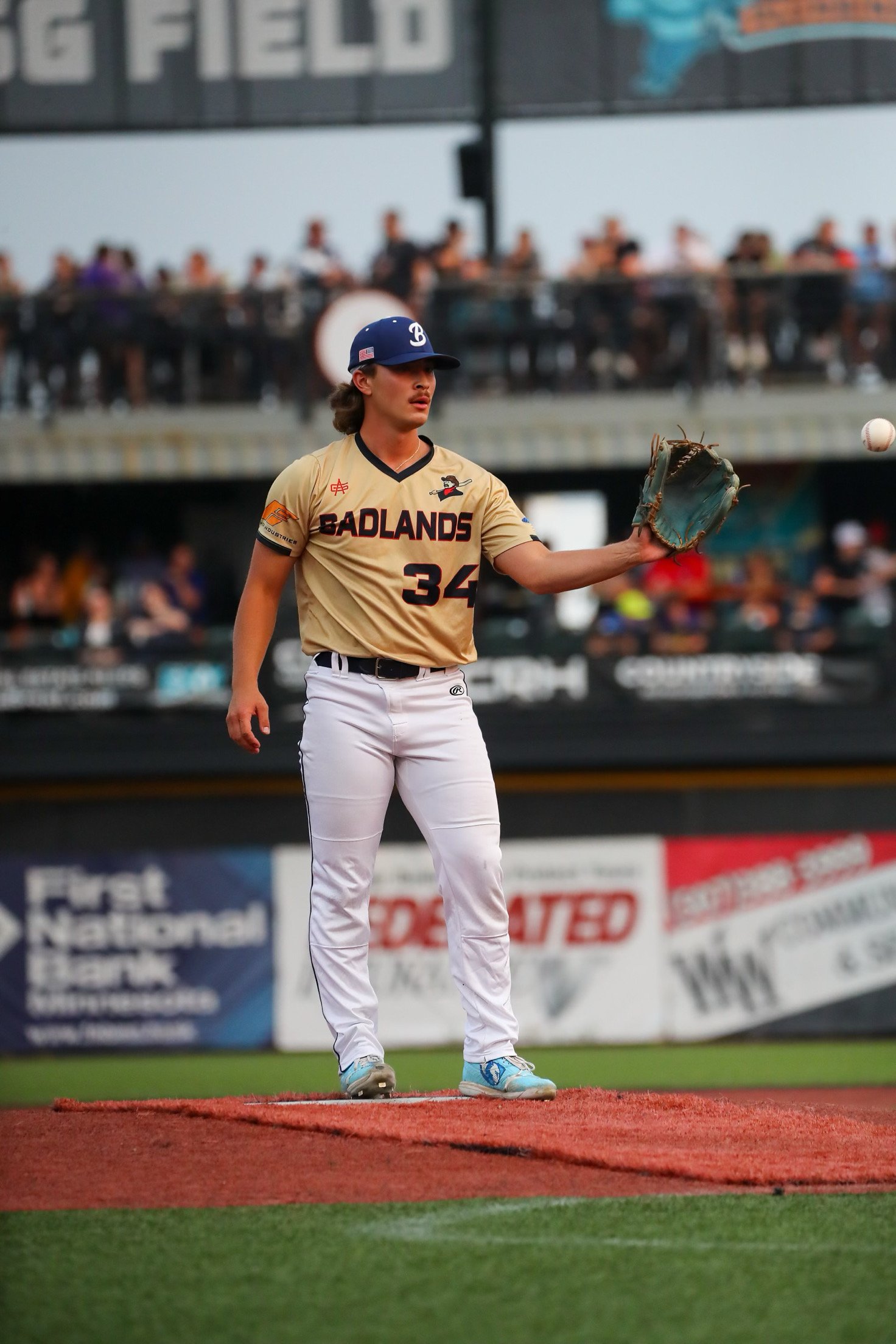A baseball player in a beige and black uniform with the word BADLANDS on it, wearing a navy blue cap, standing on a baseball field, preparing for a pitch or catch, with spectators in the background.