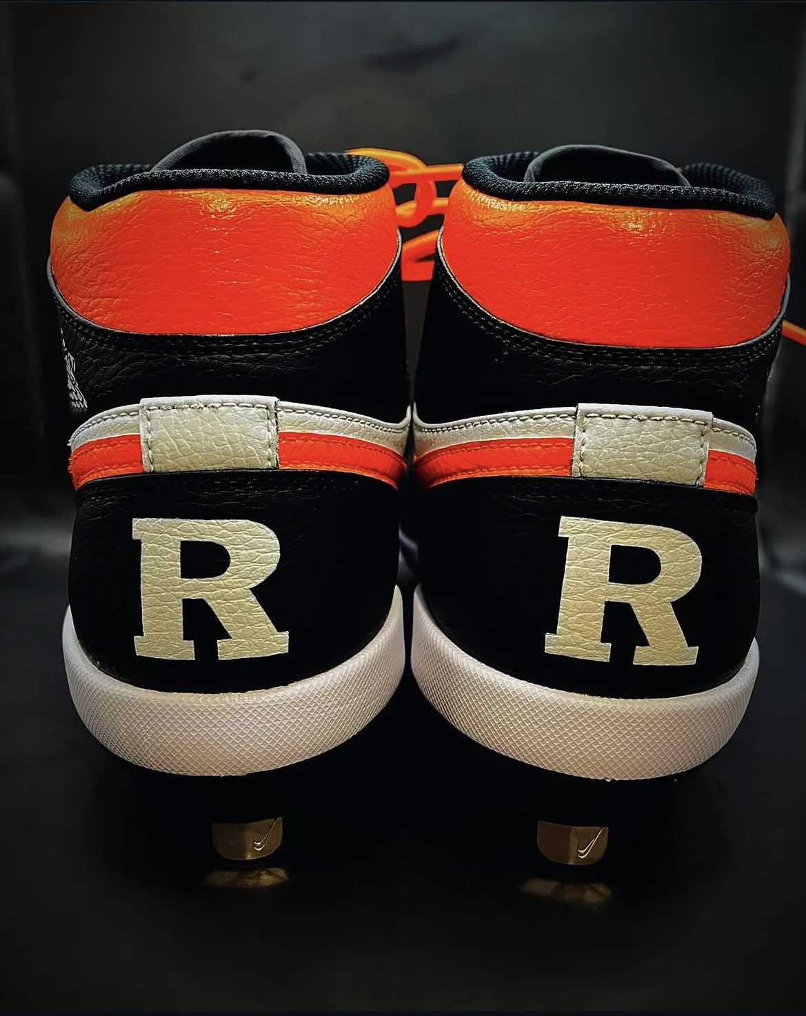 Back view of two black and orange Nike sneakers with white soles, featuring large white letter R on the back, placed on a dark surface.