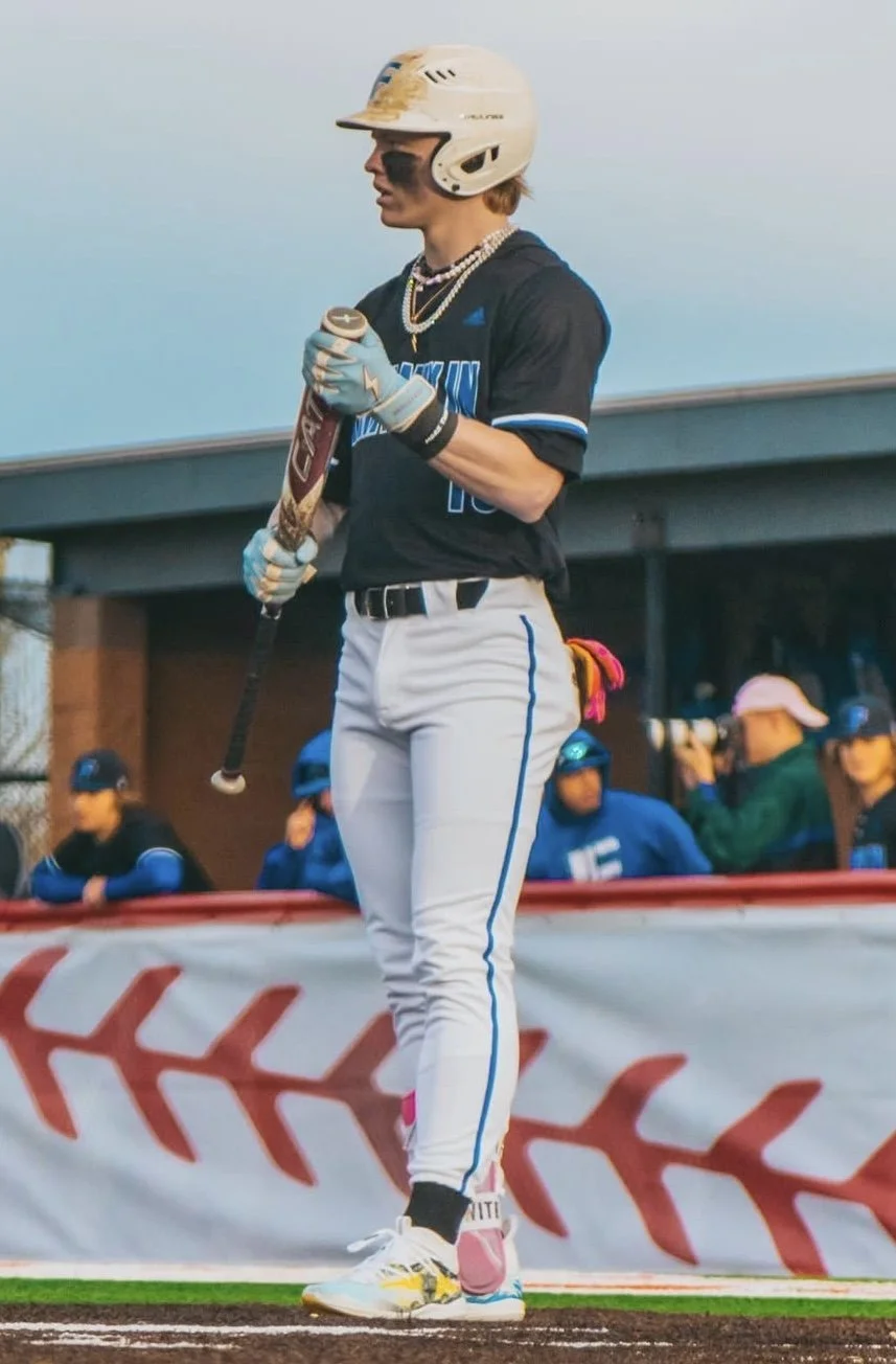 Female softball player standing on the field with a bat, wearing a helmet and uniform, with several spectators or teammates in the background.