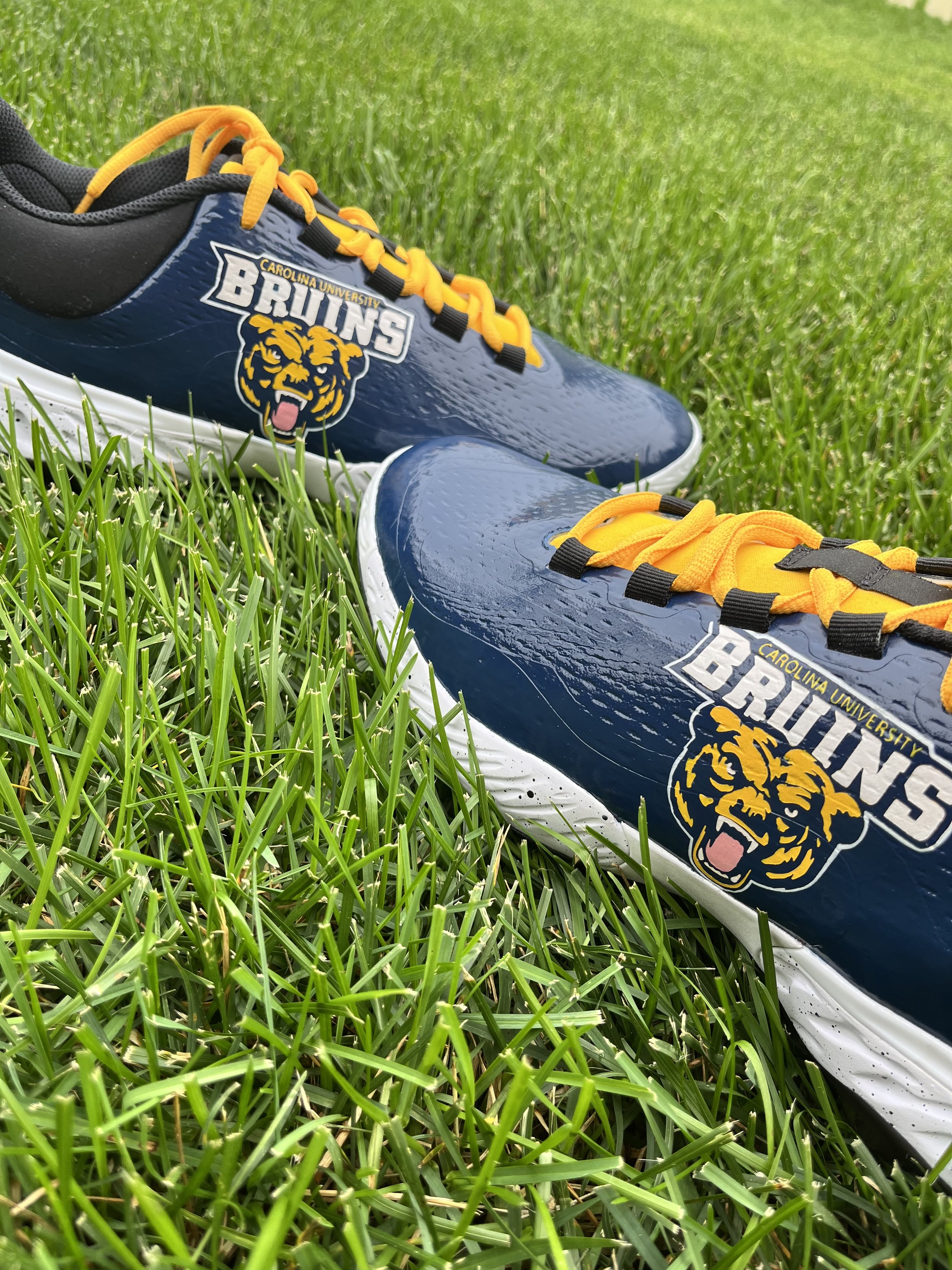 A pair of navy blue running shoes with yellow laces and a Carolina University Bruins logo on the side, lying on green grass.