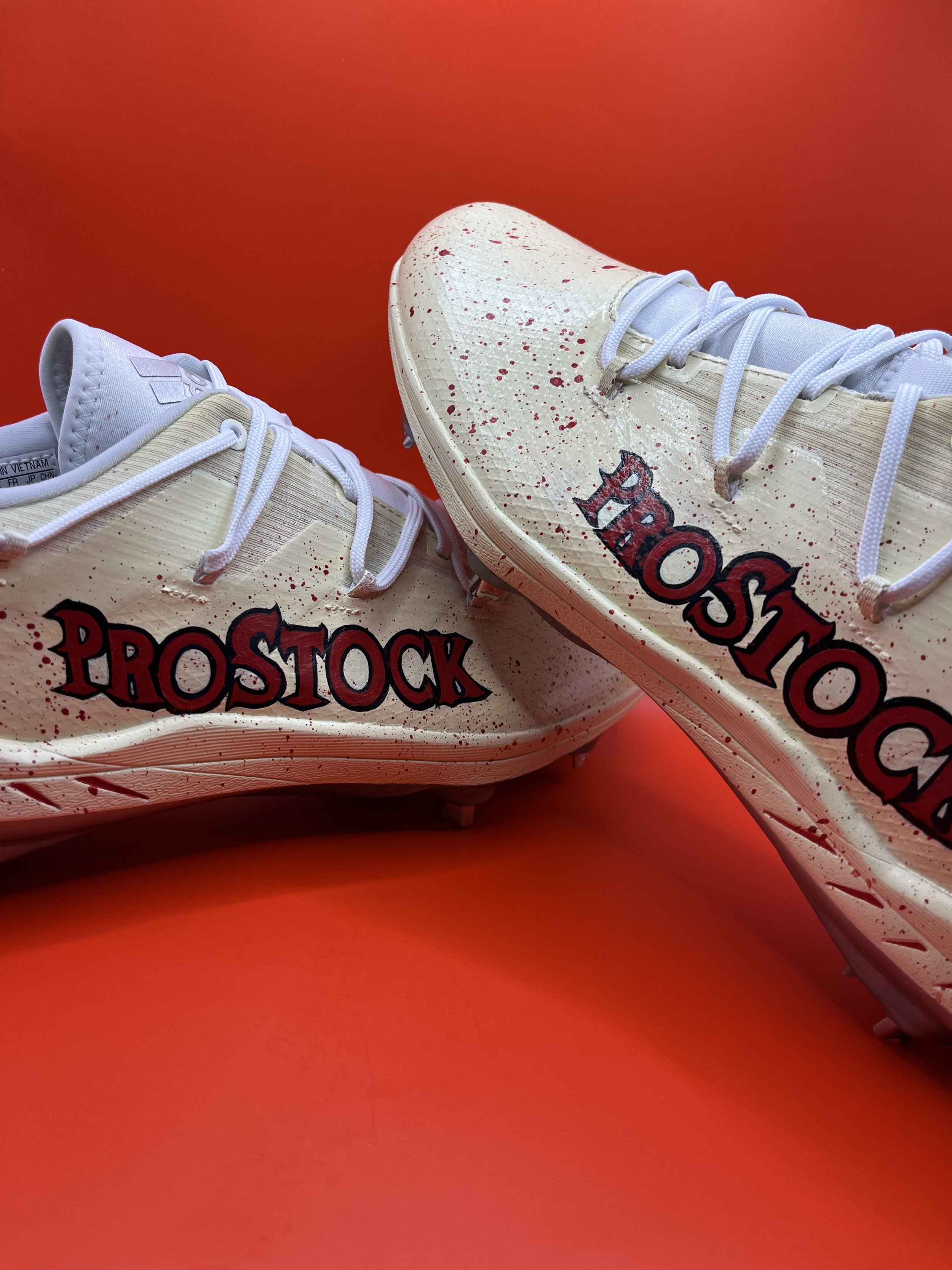 Close-up image of a pair of white soccer cleats with red paint splatter design and the word "PROSTOCK" printed on the side, set against a solid red background.