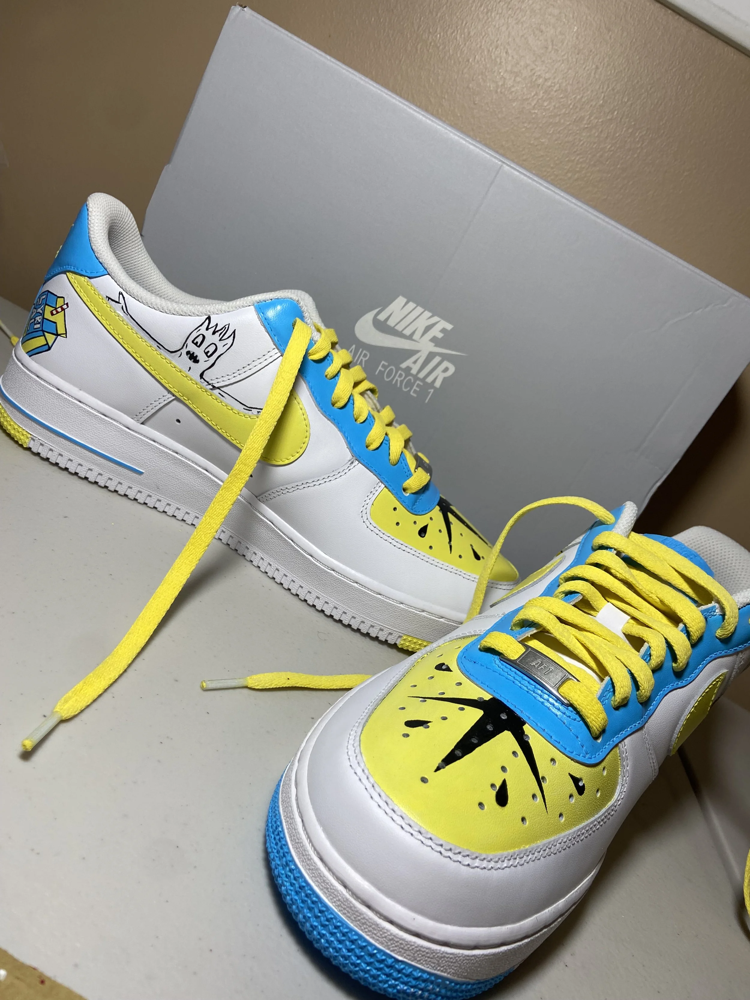 Colorful Nike Air Force 1 sneakers with yellow laces, featuring black star and hand-drawn illustrations on the sides and toe caps, placed near a Nike Air Force 1 shoe box.
